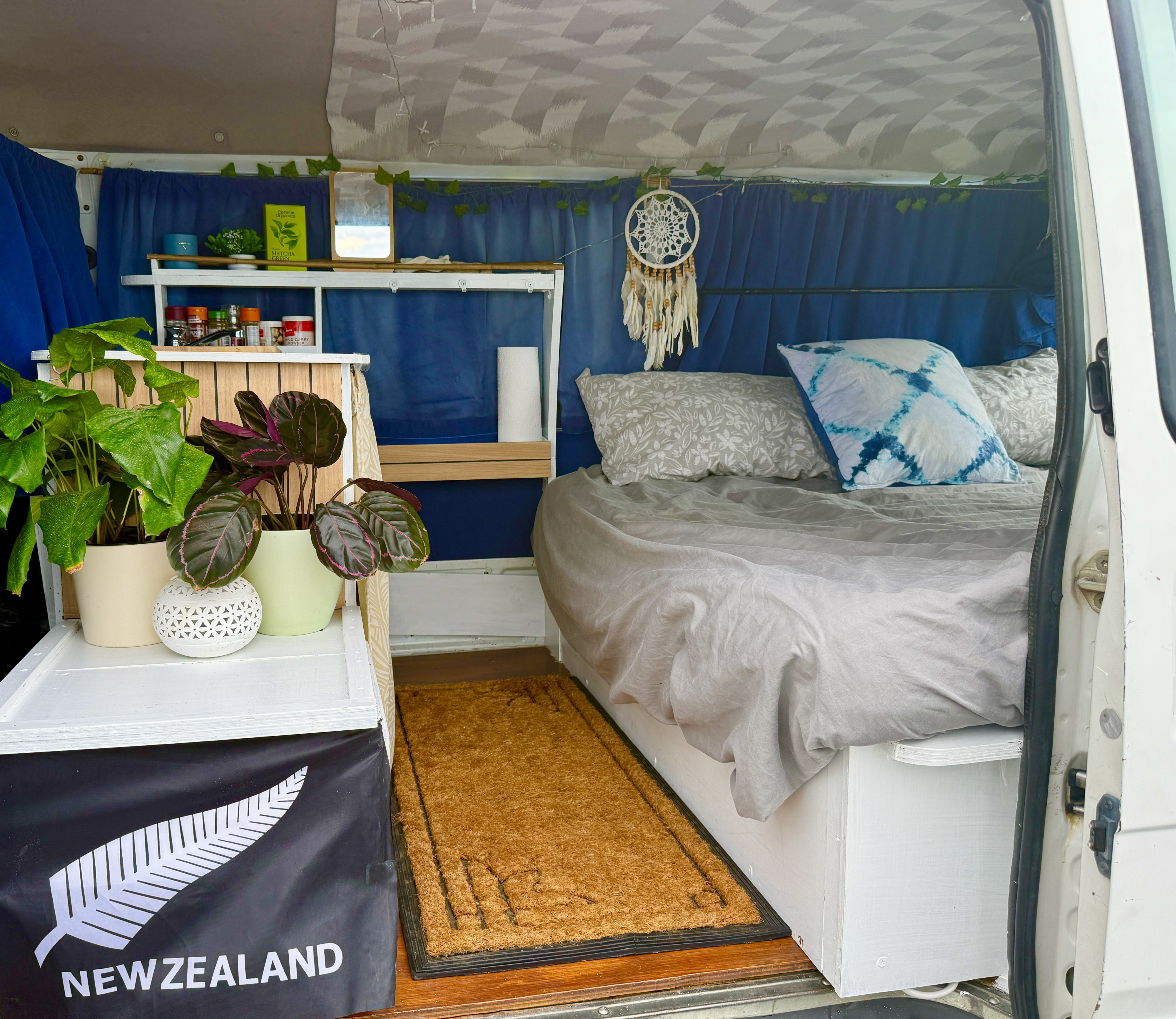 Step into this cozy RV rental interior, featuring a comfy bed, lush plants, blue curtains, and decor for a home-like travel experience.. Mazda Bongo, Gekko conversion 2004