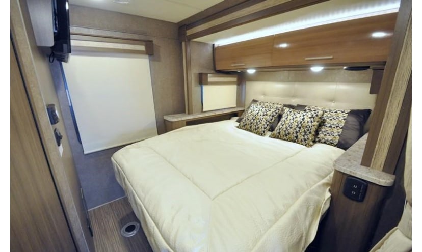 Cozy rv rental bedroom interior with a queen bed, upholstered headboard, overhead cabinets, and a window with a shade.. Thor Gemini 2018