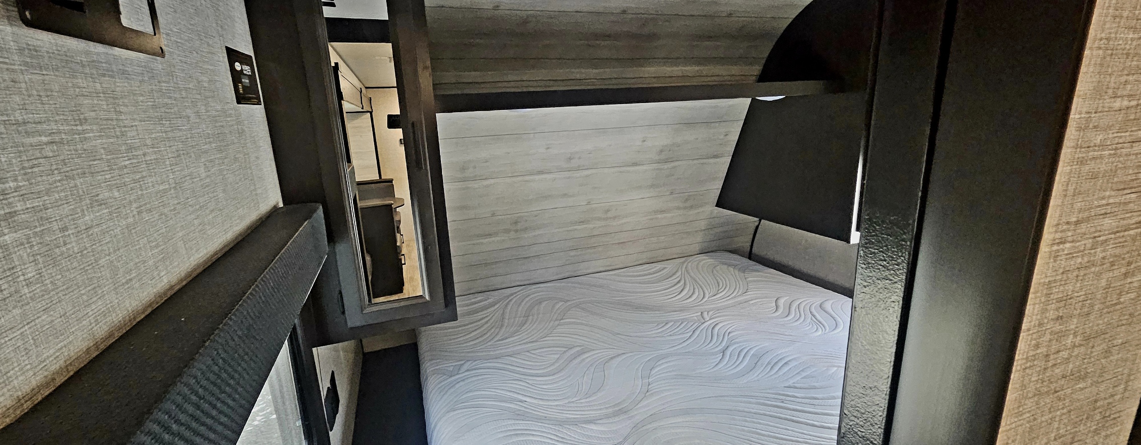 Explore this RV rental bedroom: a modern space featuring a patterned white mattress, wood-style wall, lamp, and overhead storage.. Jayco Jay Flight SLX 2022