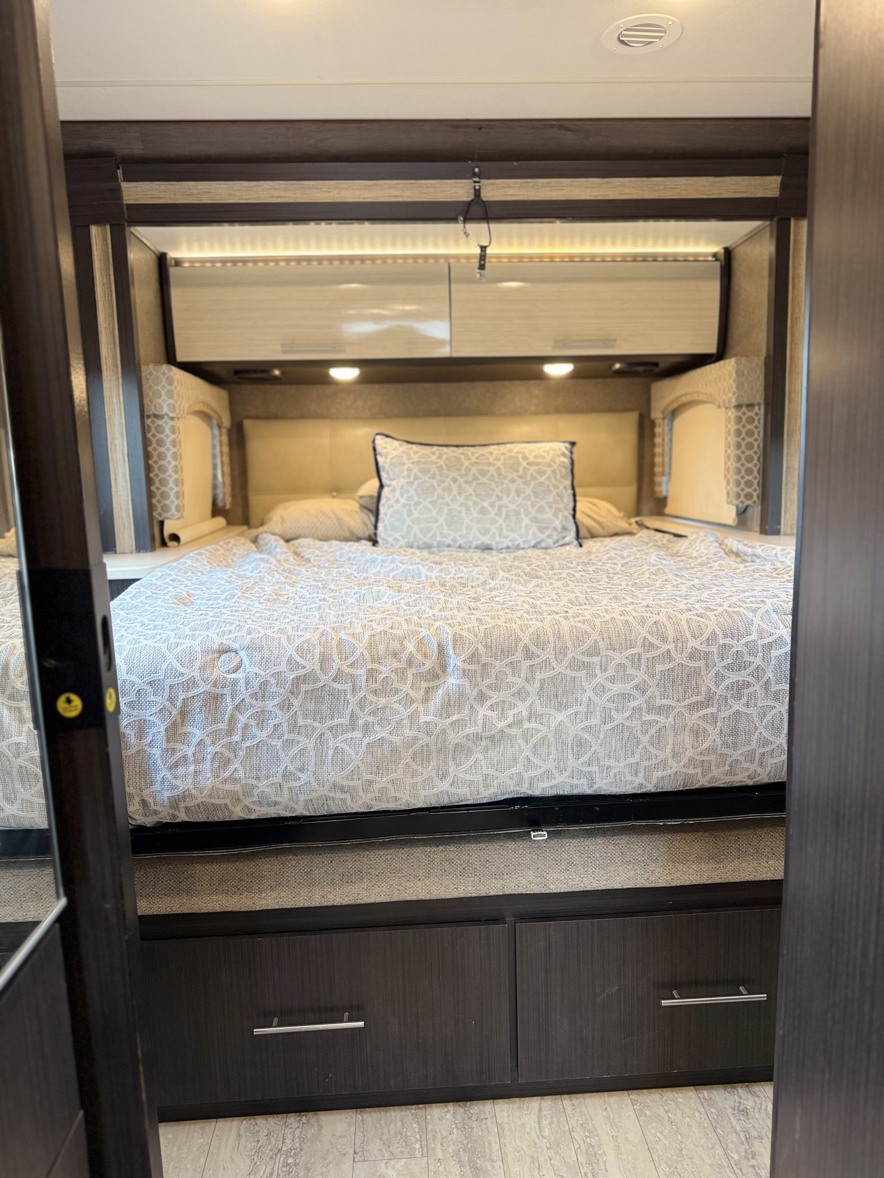 Comfortable RV rental bedroom featuring a bed with patterned bedding, overhead storage, ambient lighting, and built-in drawers. Ideal for travel.. Thor Axis 2018