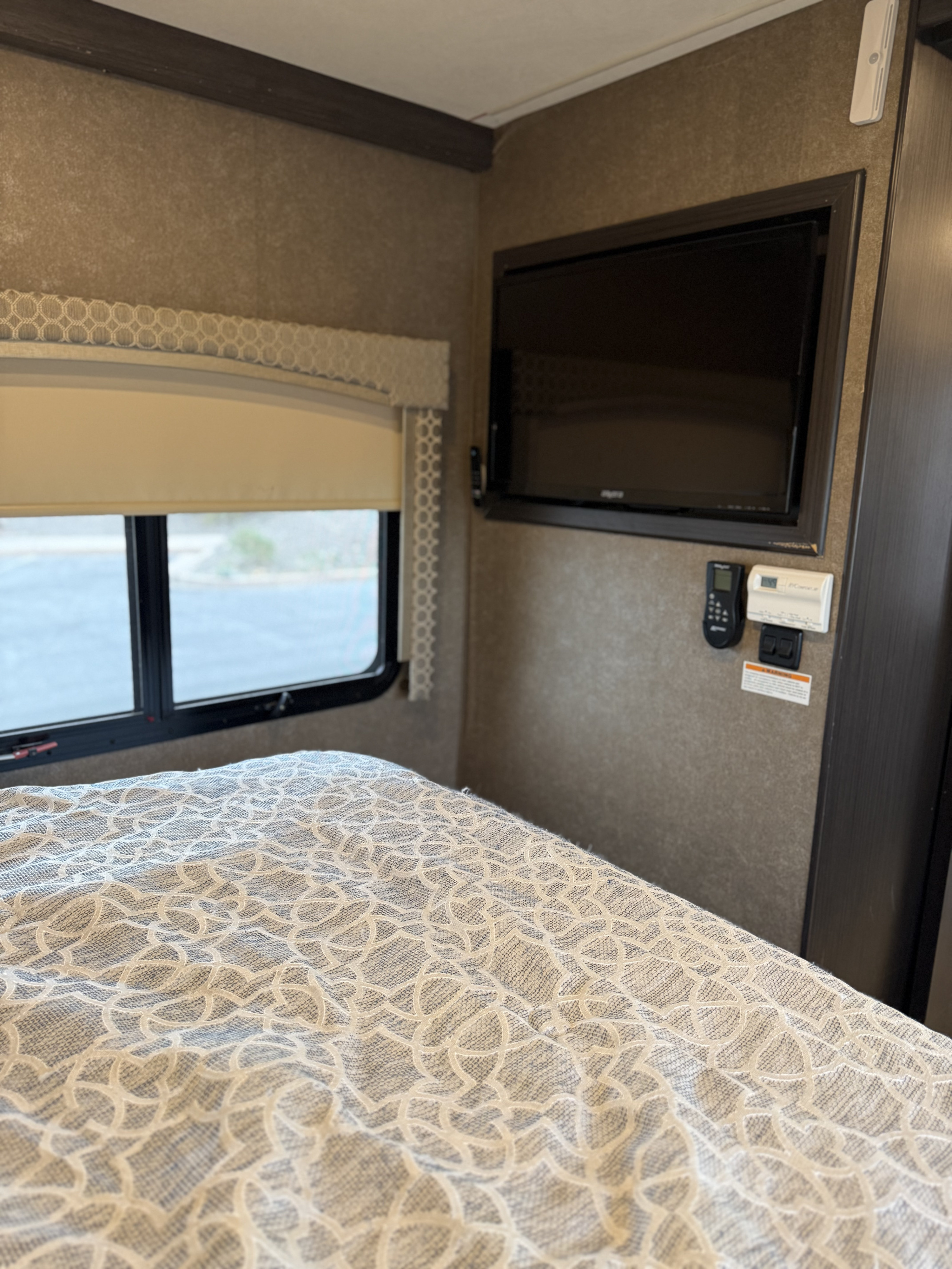 Cozy RV rental bedroom with a patterned bedspread, wall-mounted TV, window view, and climate controls on the right.. Thor Axis 2018