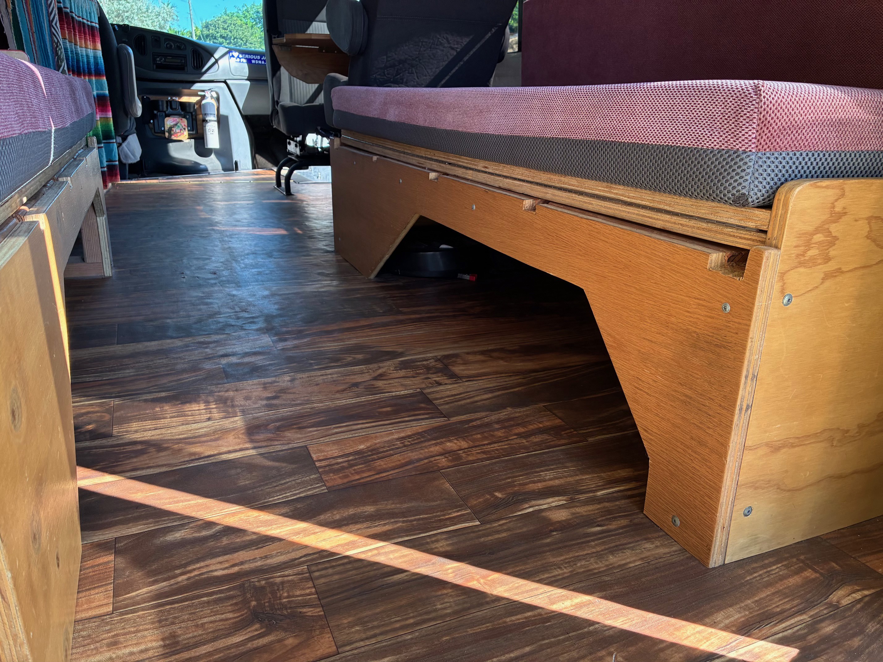 Rv rental interior: wood plank floor, custom wooden bench/bed frame with pink and grey cushion. Van cockpit in sunlit space.. Ford E350 Box Van 2004