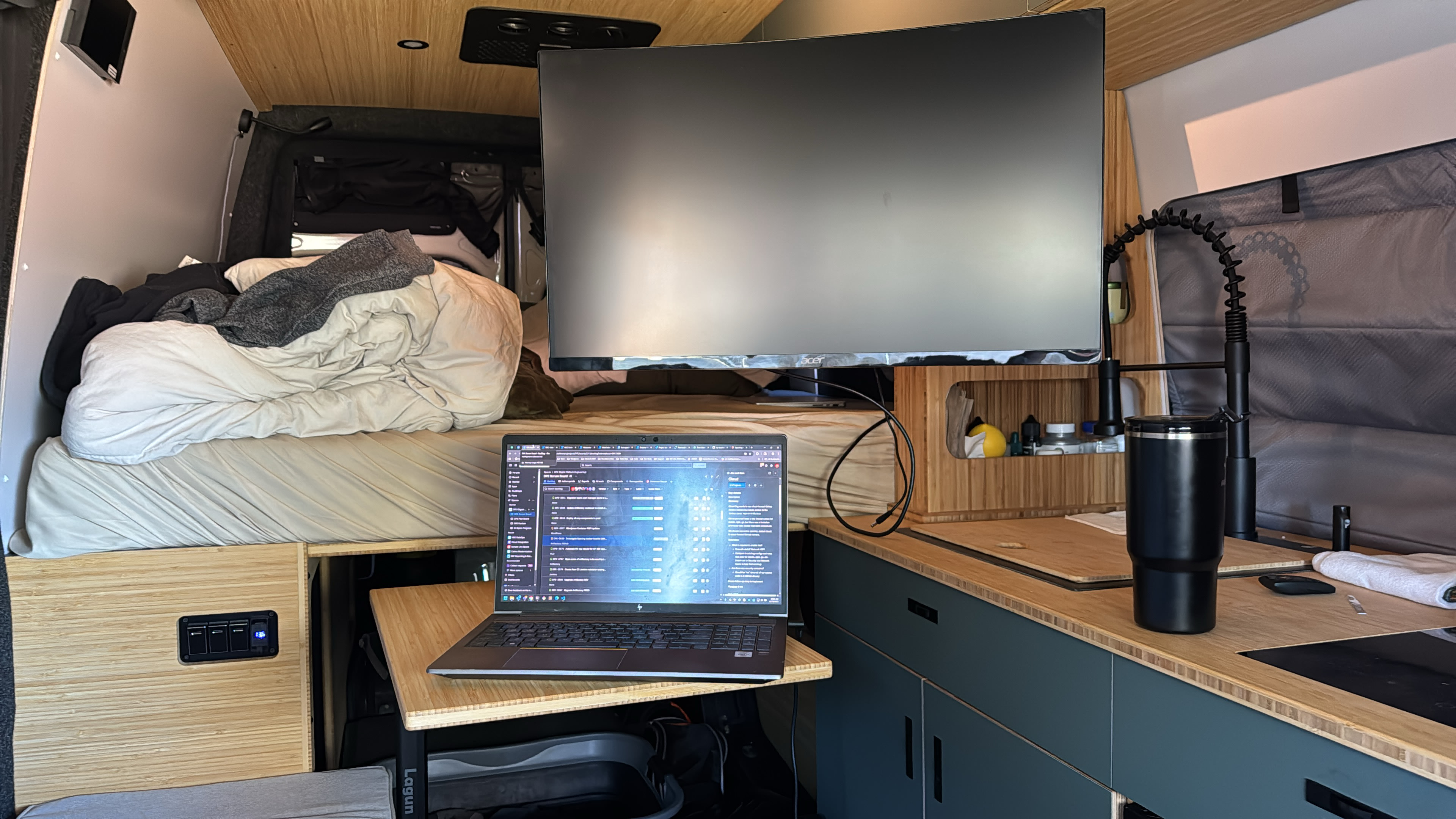 Modern RV rental interior: large monitor, laptop workstation, cozy bed, and fully equipped kitchen area for mobile living.. Mercedes Sprinter 2025