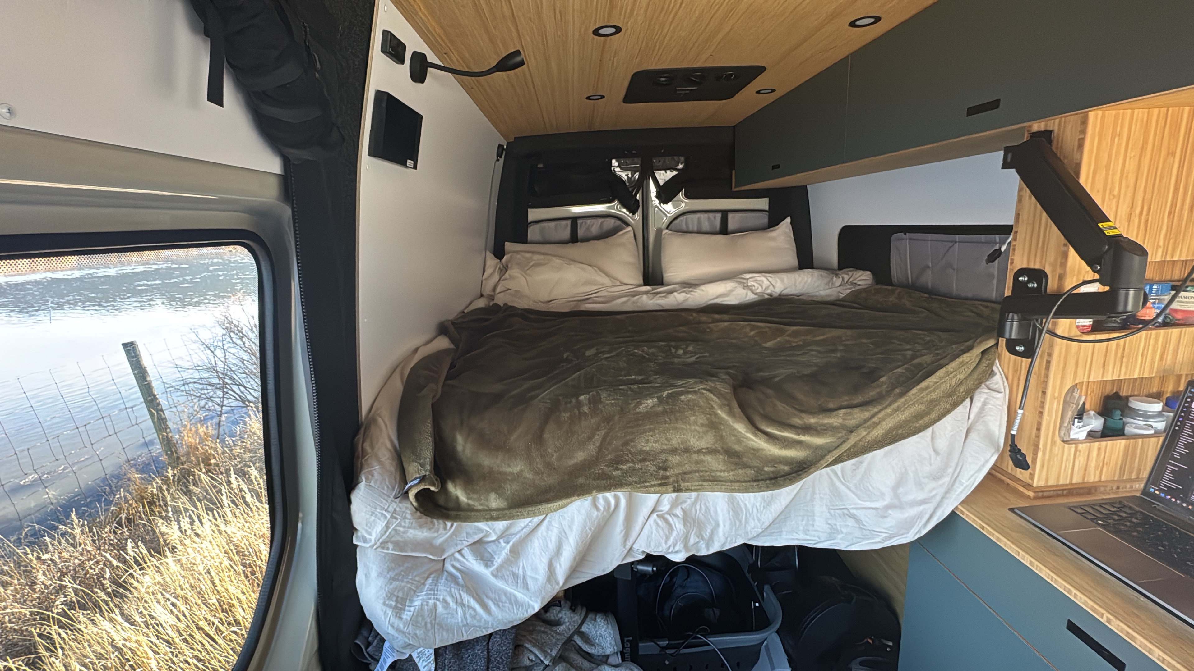 Cozy rv rental interior: bed with olive blanket, laptop workspace, and river view. Ideal for van life travel.. Mercedes Sprinter 2025