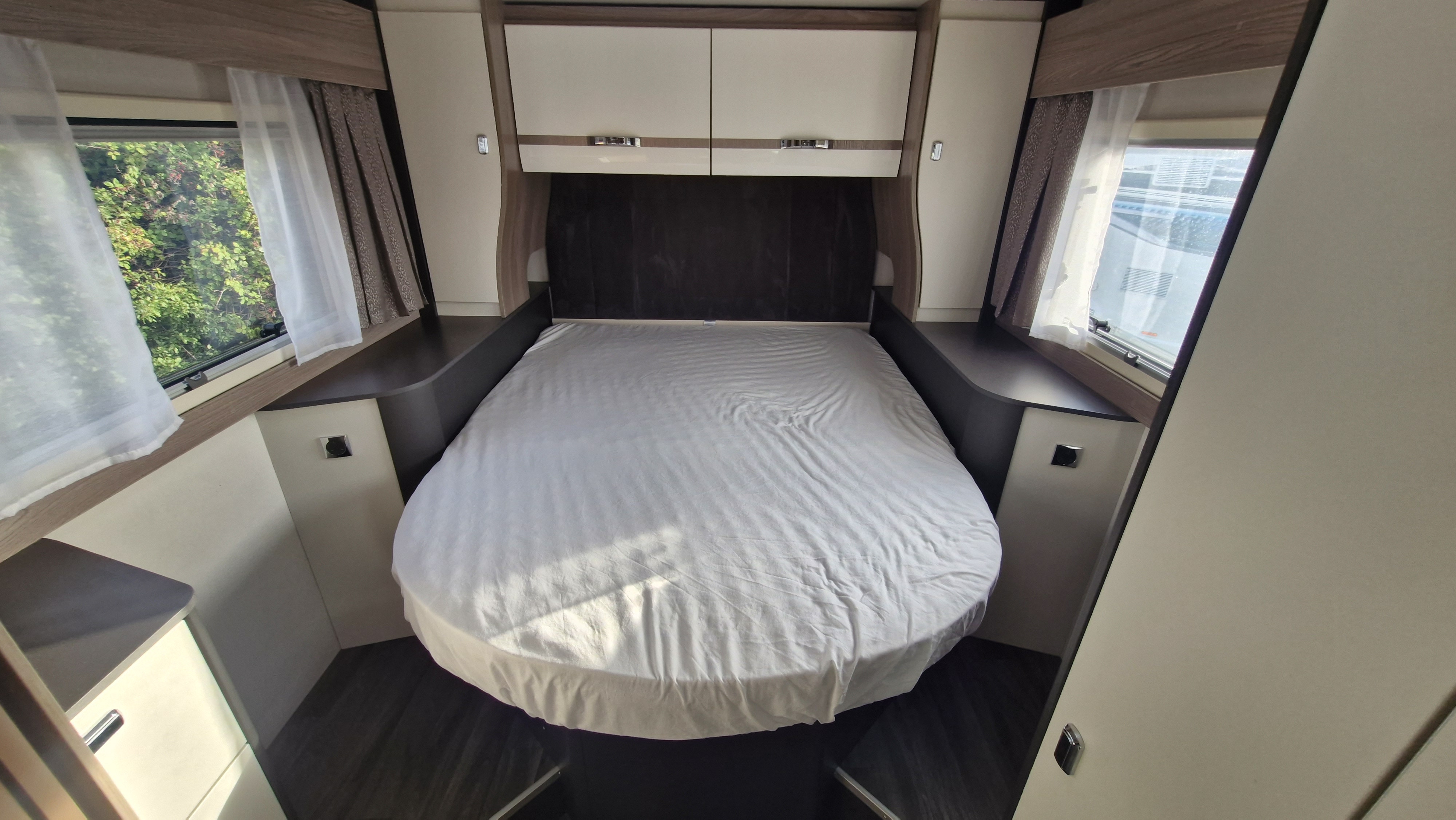 Cozy RV rental bedroom featuring a large bed with white sheets, overhead cabinets, and windows with curtains for scenic views.. Swift Voyager 494 2026