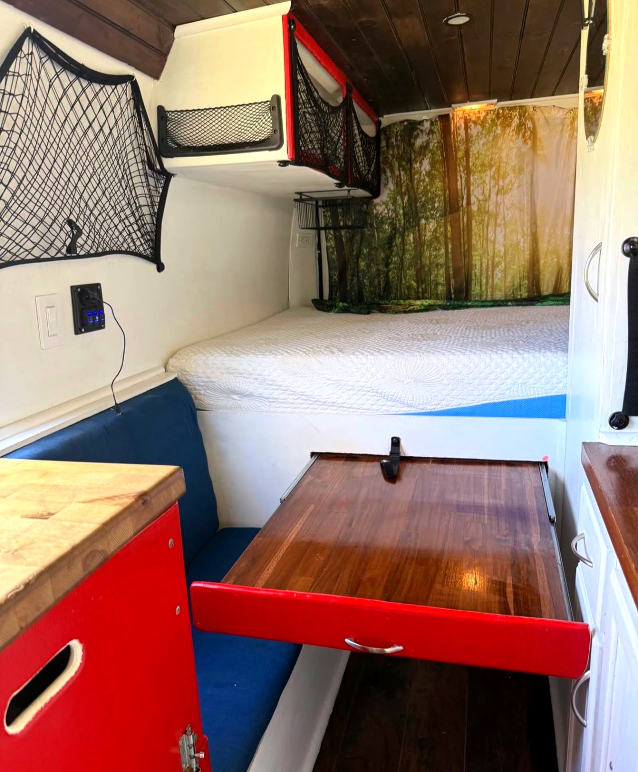 Inside this RV rental: a bed, pull-out wooden table, blue bench, storage nets, and a forest curtain.. Ram Promaster 2500 2019