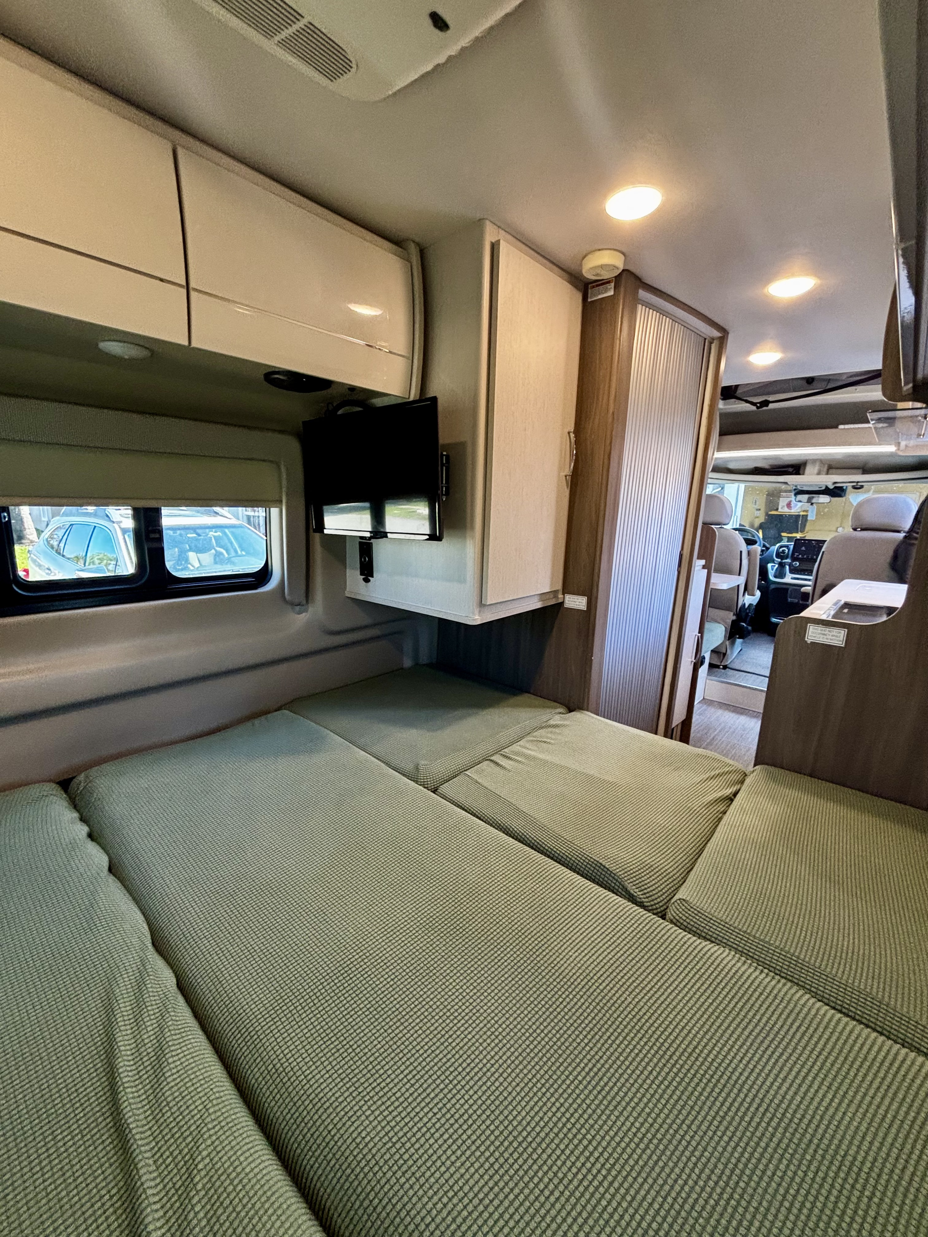 Cozy RV rental interior: green cushioned bed, windows, TV, overhead storage, and a view of the front cabin.. Thor Tellaro 2023