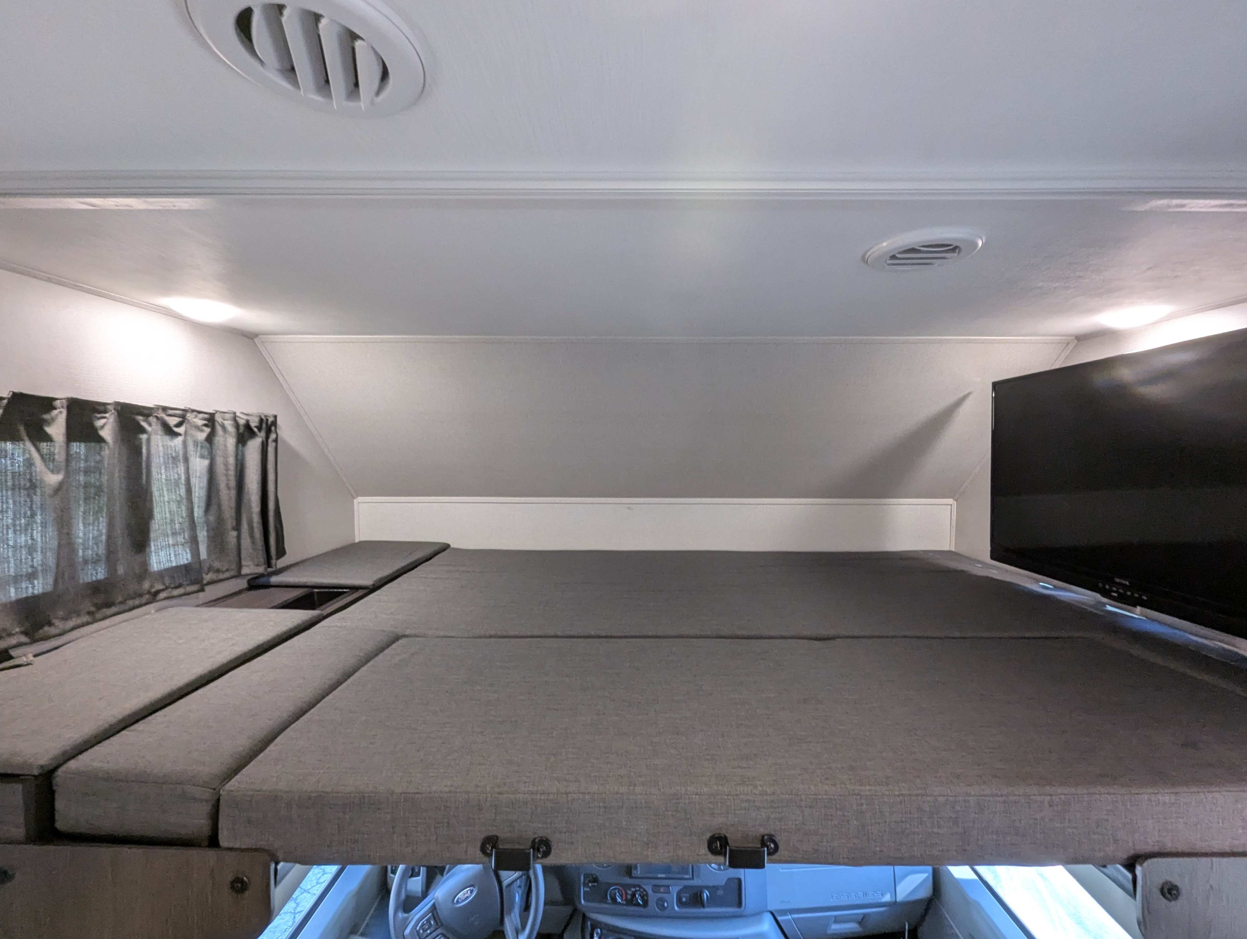 Inside an RV rental, a cozy cab-over bunk bed with cushions, a window with curtains, and a mounted TV.. Forest River Forester 2023