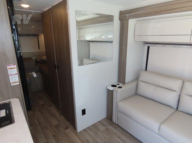 Bright RV rental living space with light sofa, overhead AC, mirror, leading to a compact bedroom and kitchen area.. Thor Axis 2024