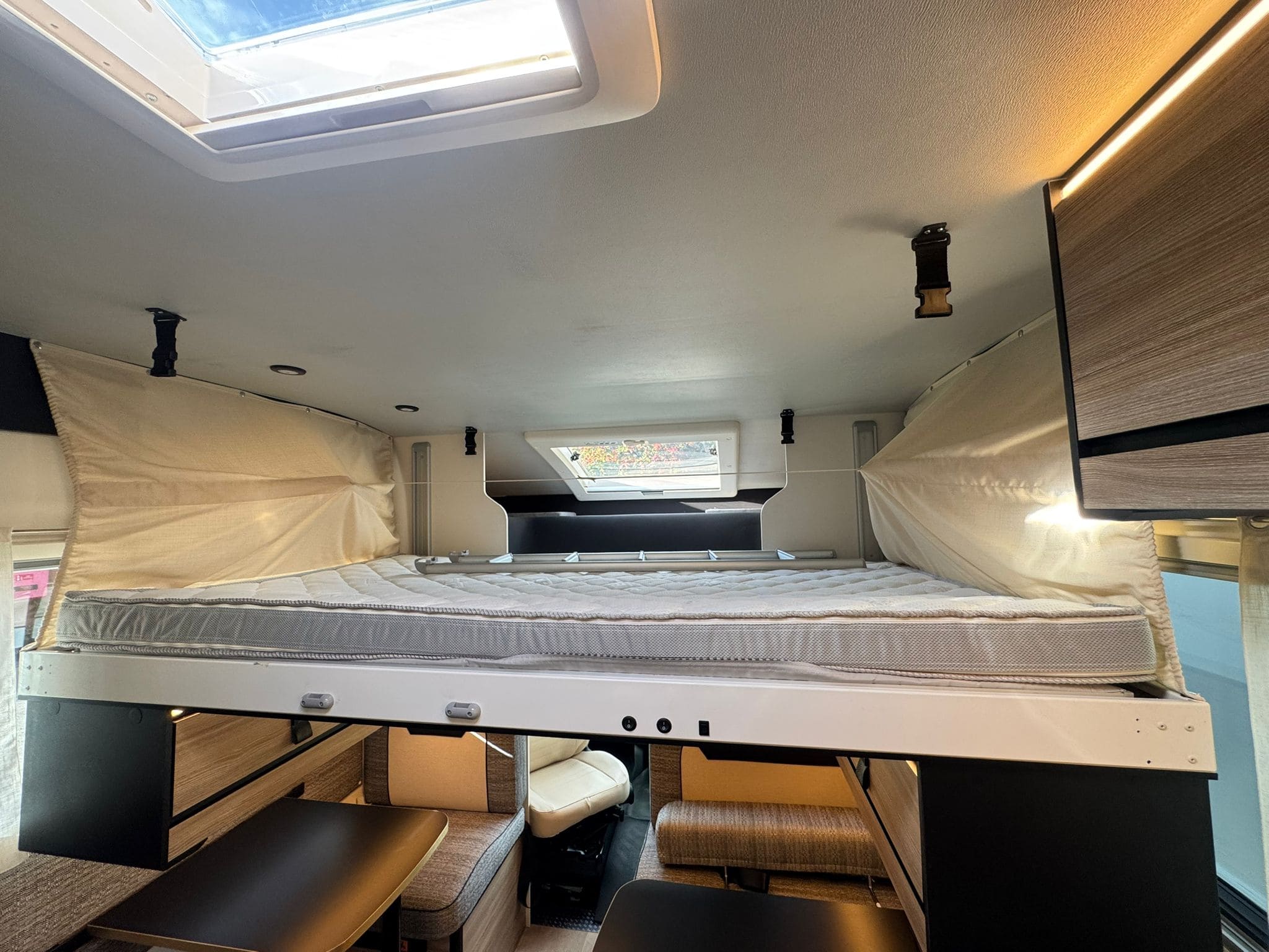 Inside an rv rental: an overhead bunk bed with a mattress, positioned above a seating area, illuminated by a skylight and ceiling lights.. FORD TRANSIT RIMOR KILIG 79+ 2026