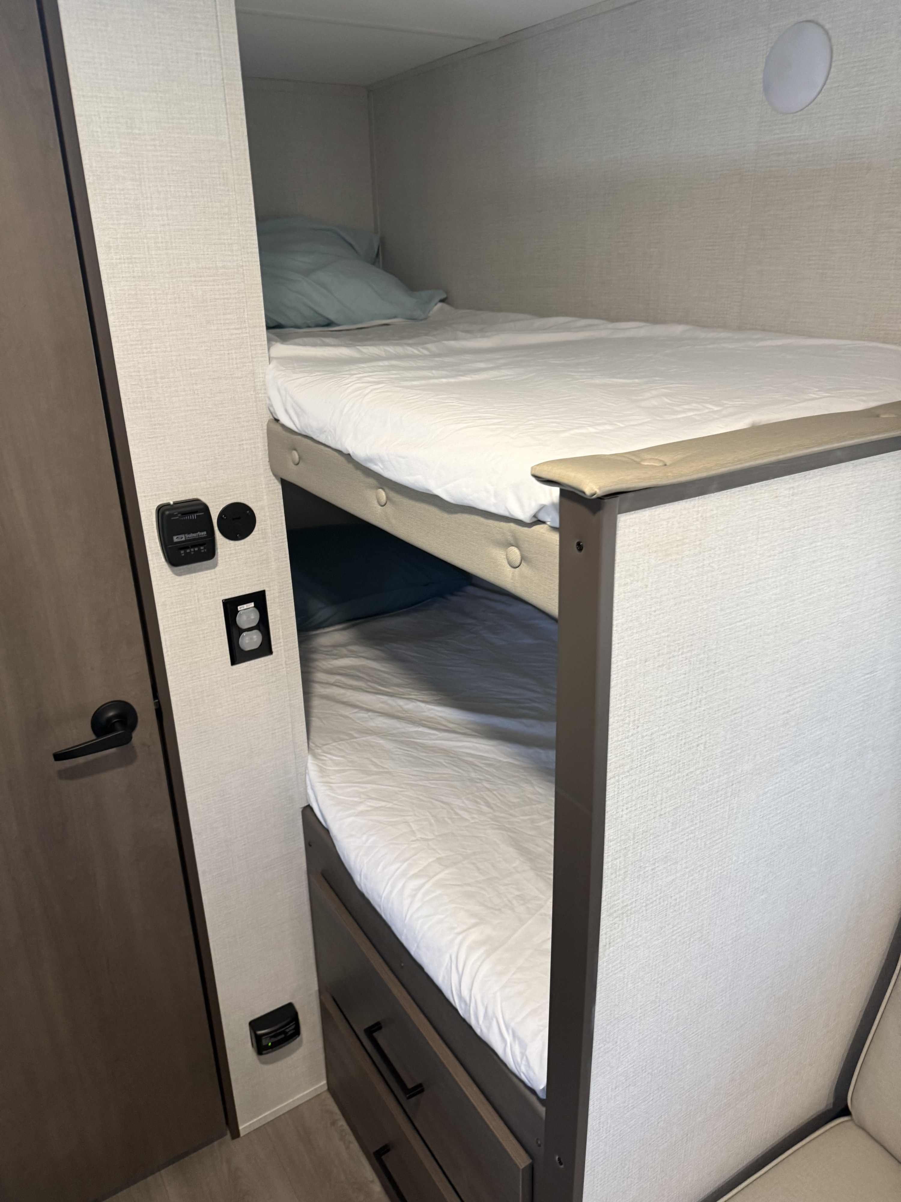 Inside an RV rental: view of stacked bunk beds with white sheets and pillows, integrated control panel and power outlets.. Grand Design Transcend One 2025