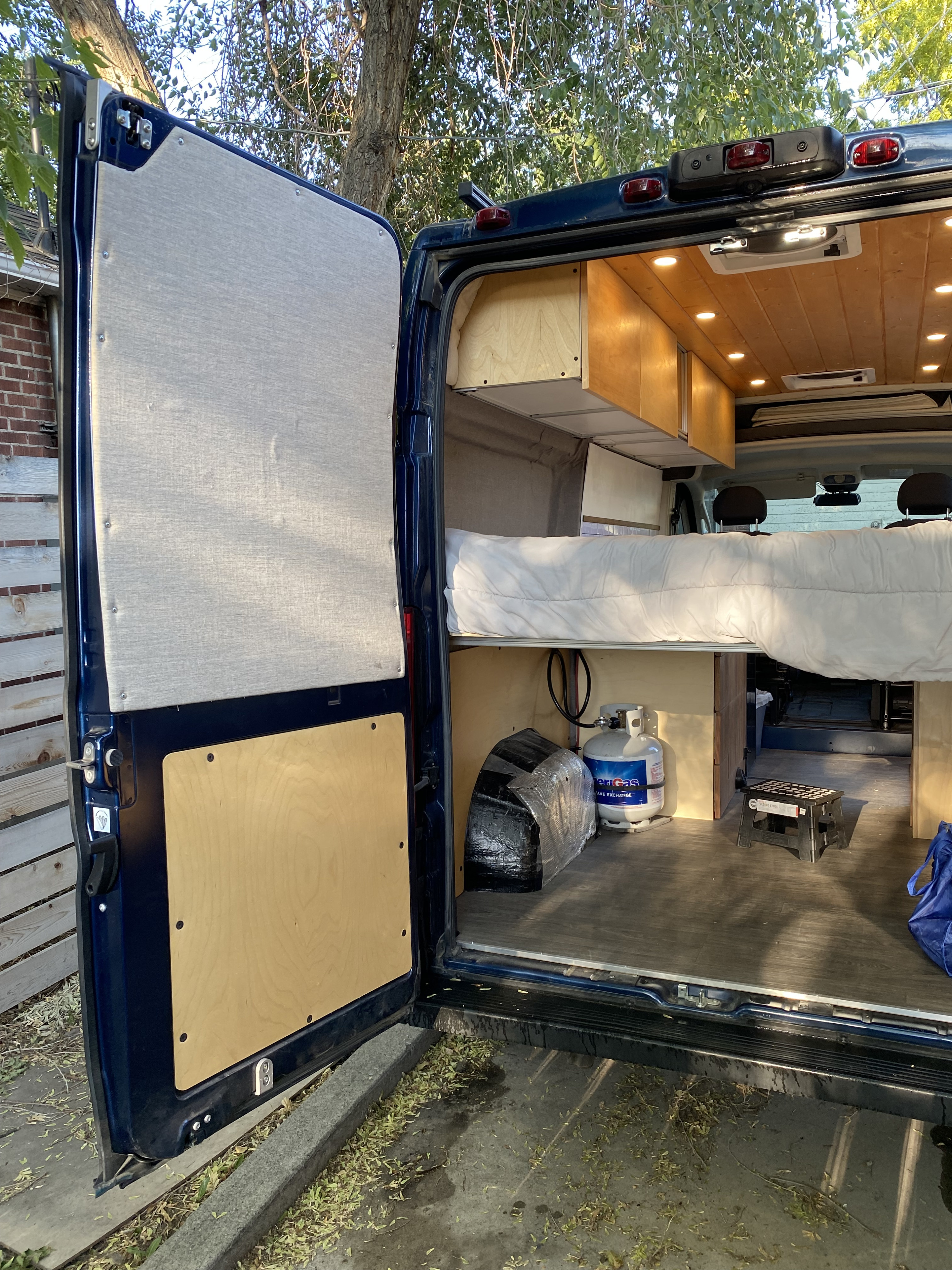 View inside an rv rental camper van, featuring a cozy bed, overhead wooden cabinets, a propane tank, and under-bed storage.. Custom Camper 2021