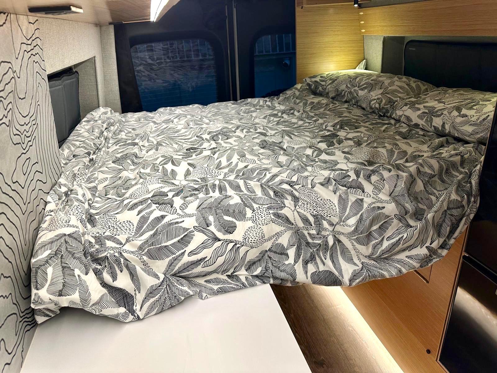 A plush bed with black/white leaf duvet inside a cozy RV rental. Features accent lighting & modern wood paneling.. Rossmonster Skye 2025