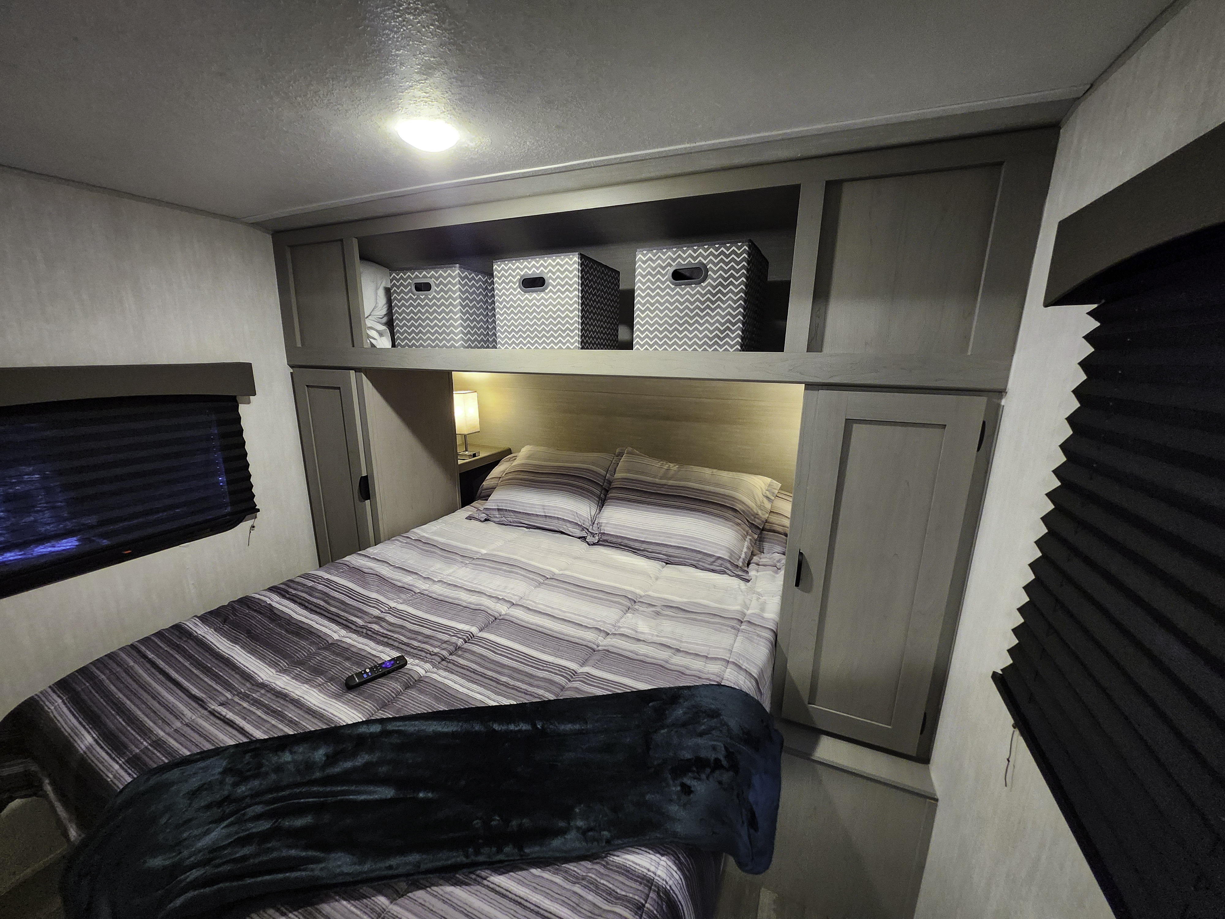 Interior of an RV rental bedroom with striped bedding, overhead storage, side windows, and a cozy lamp for comfort.. Forest River Viking 6K 2025