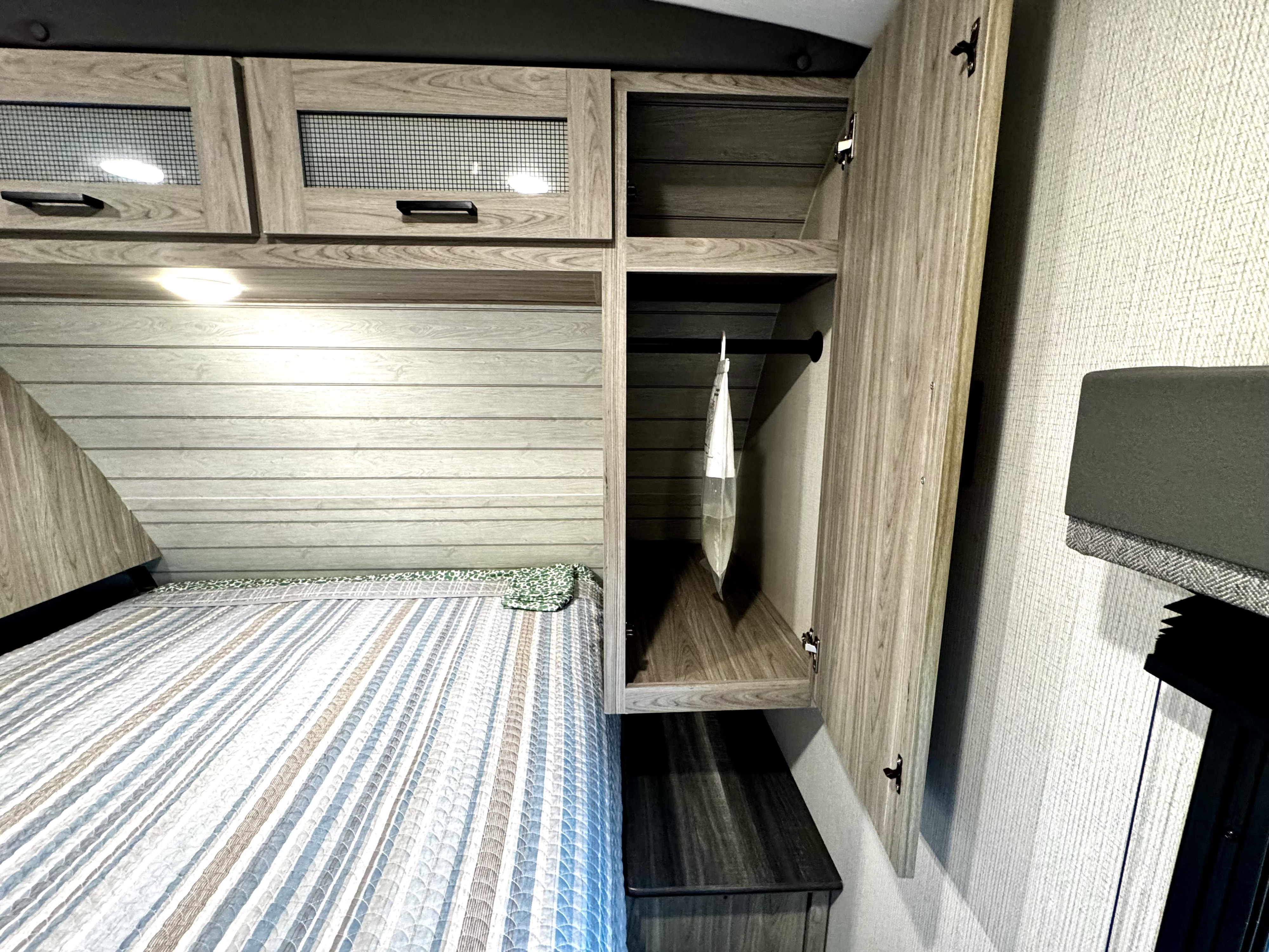 Cozy RV rental bedroom with a striped bed, overhead cabinets, and an open wardrobe featuring a hanging bar for storage.. Keystone RV Passport GT 2022
