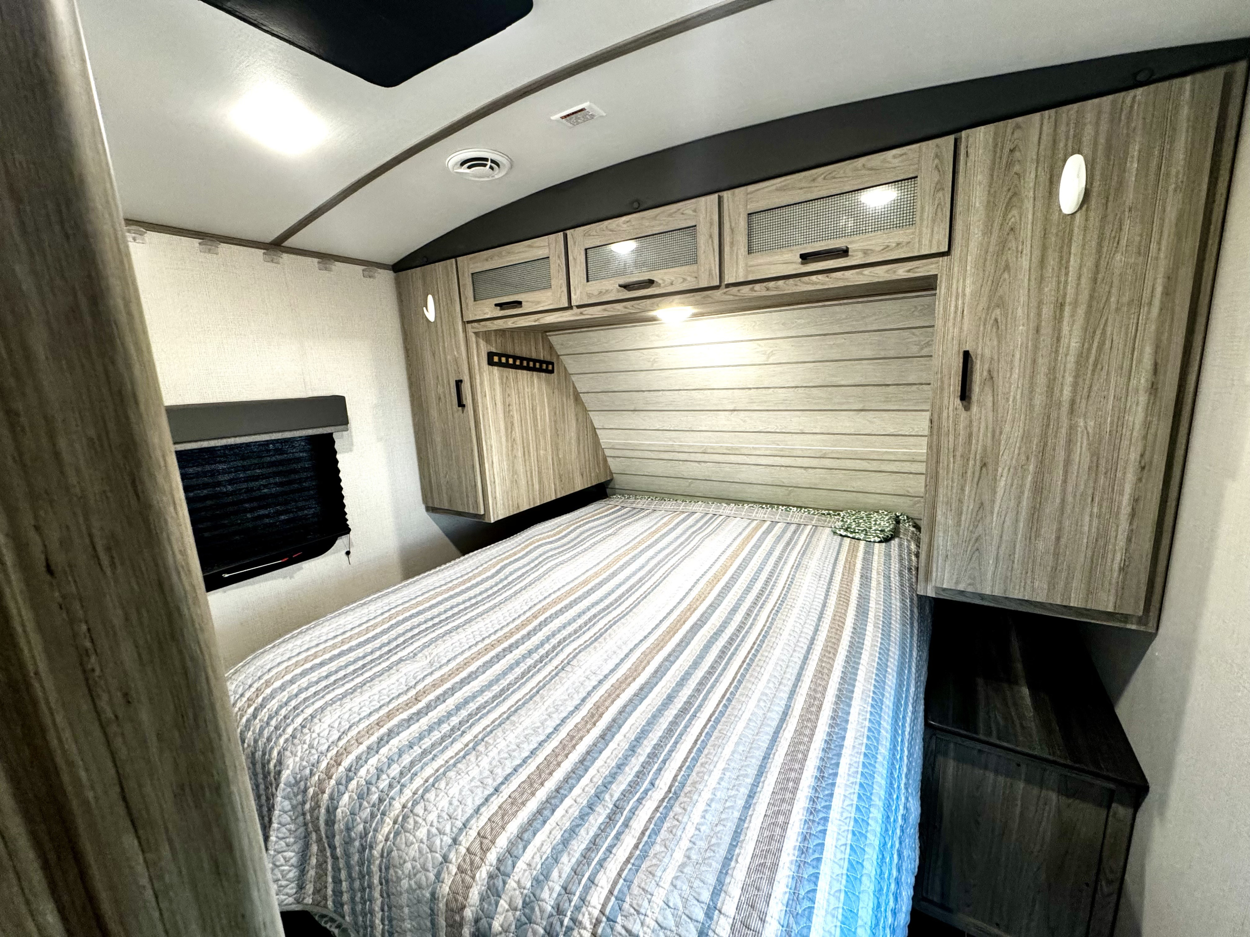 Cozy RV rental bedroom featuring a striped bed, overhead storage cabinets, and a window with a blackout blind.. Keystone RV Passport GT 2022