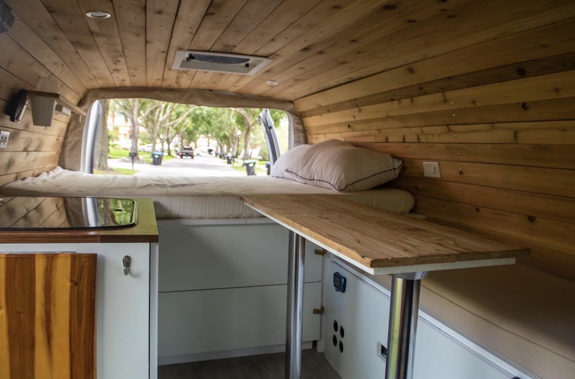 Interior of an RV rental with wooden paneling, a bed, kitchen counter, and a table. View of a street outside.. Ford Econoline 150 2010
