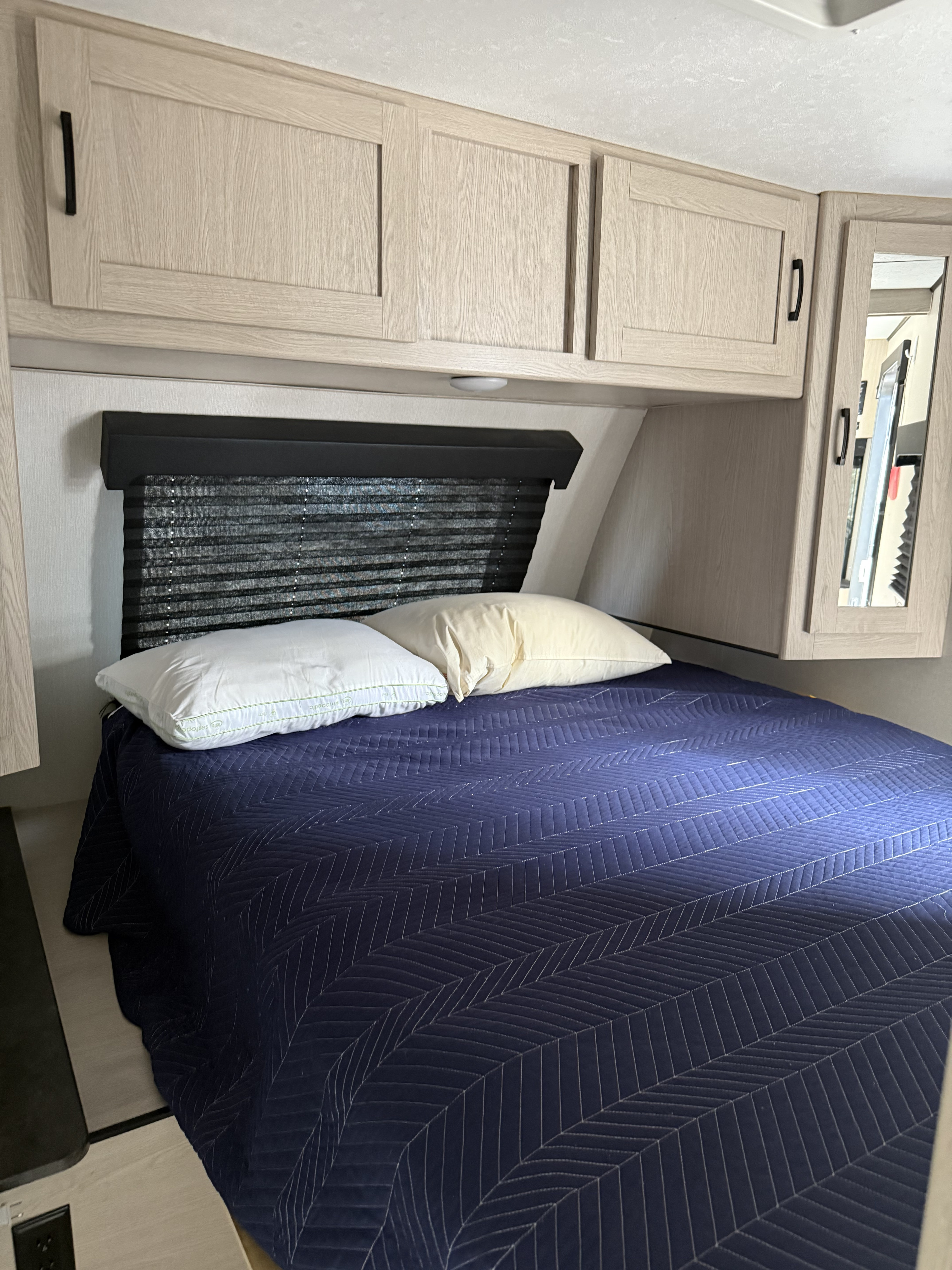 Cozy RV rental bedroom featuring light wood cabinets, a blue quilted bedspread, two pillows, and pleated window blinds.. Coachmen Apex 2021
