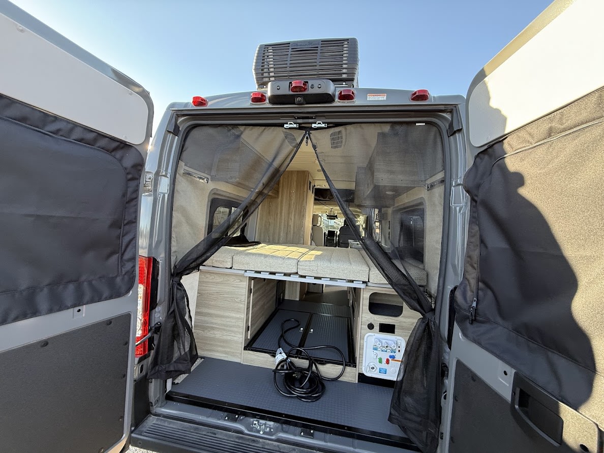 Inside an RV rental camper van, showing open rear doors, a bed, storage, control panel, and protective mesh screen.. Winnebago Solis 59PX 2026