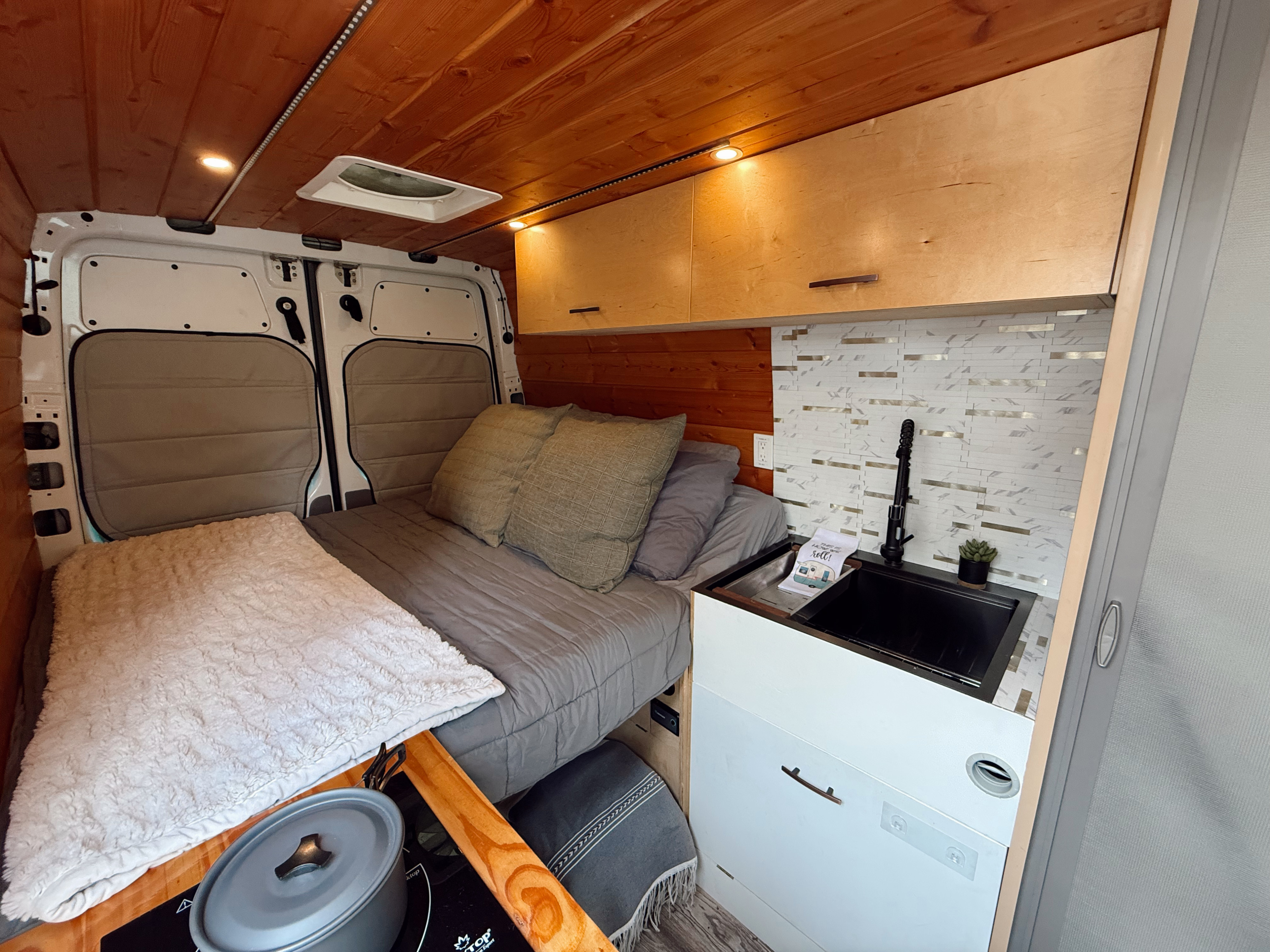 Cozy RV rental interior featuring a comfortable bed, modern kitchenette with sink and backsplash, wood-paneled ceiling, and storage.. Mercedes-Benz Sprinter 2500 2014
