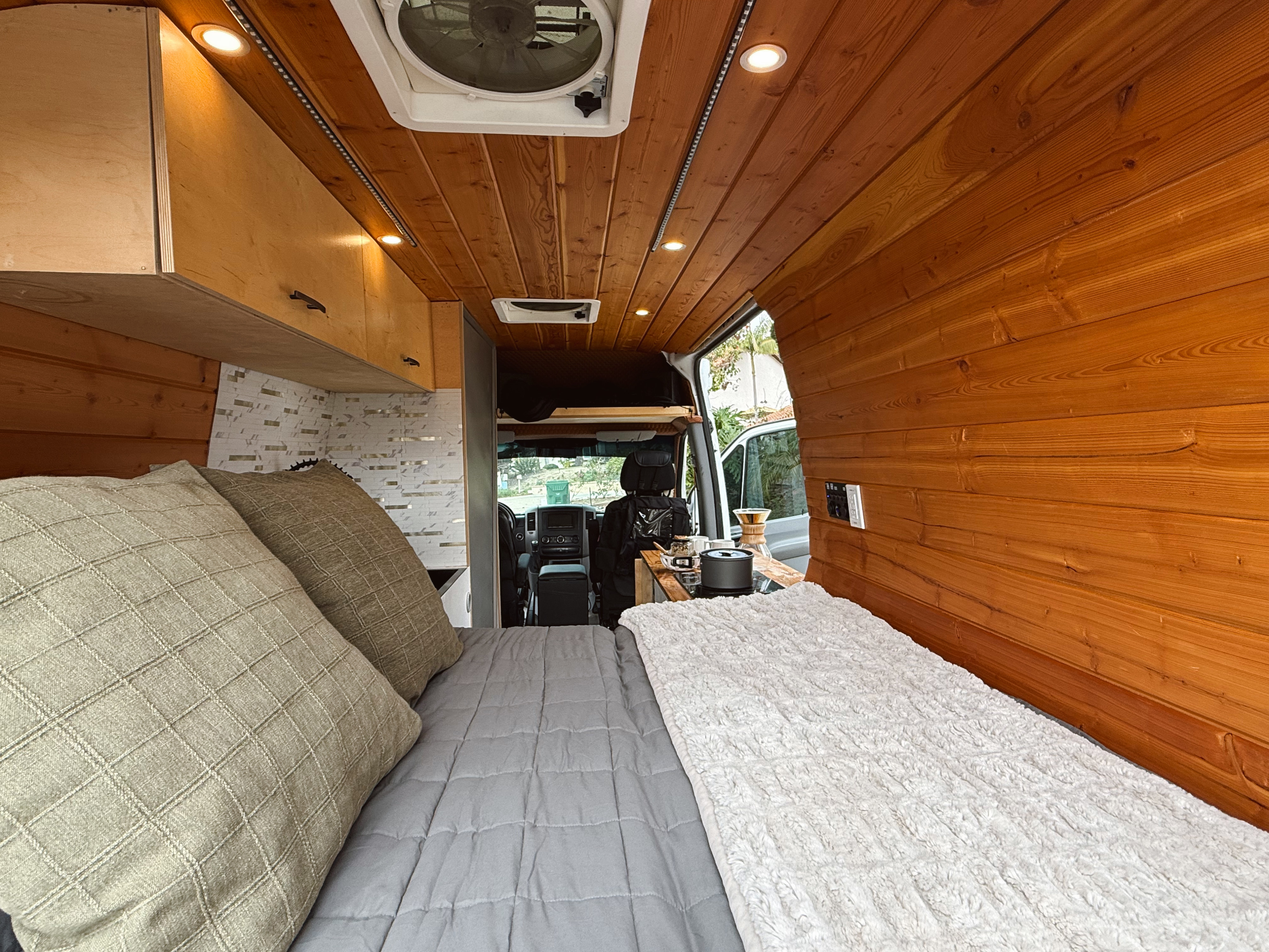Cozy RV rental interior with a comfy bed, warm wood paneling, and a compact kitchen. View extends to the driver's cab.. Mercedes-Benz Sprinter 2500 2014