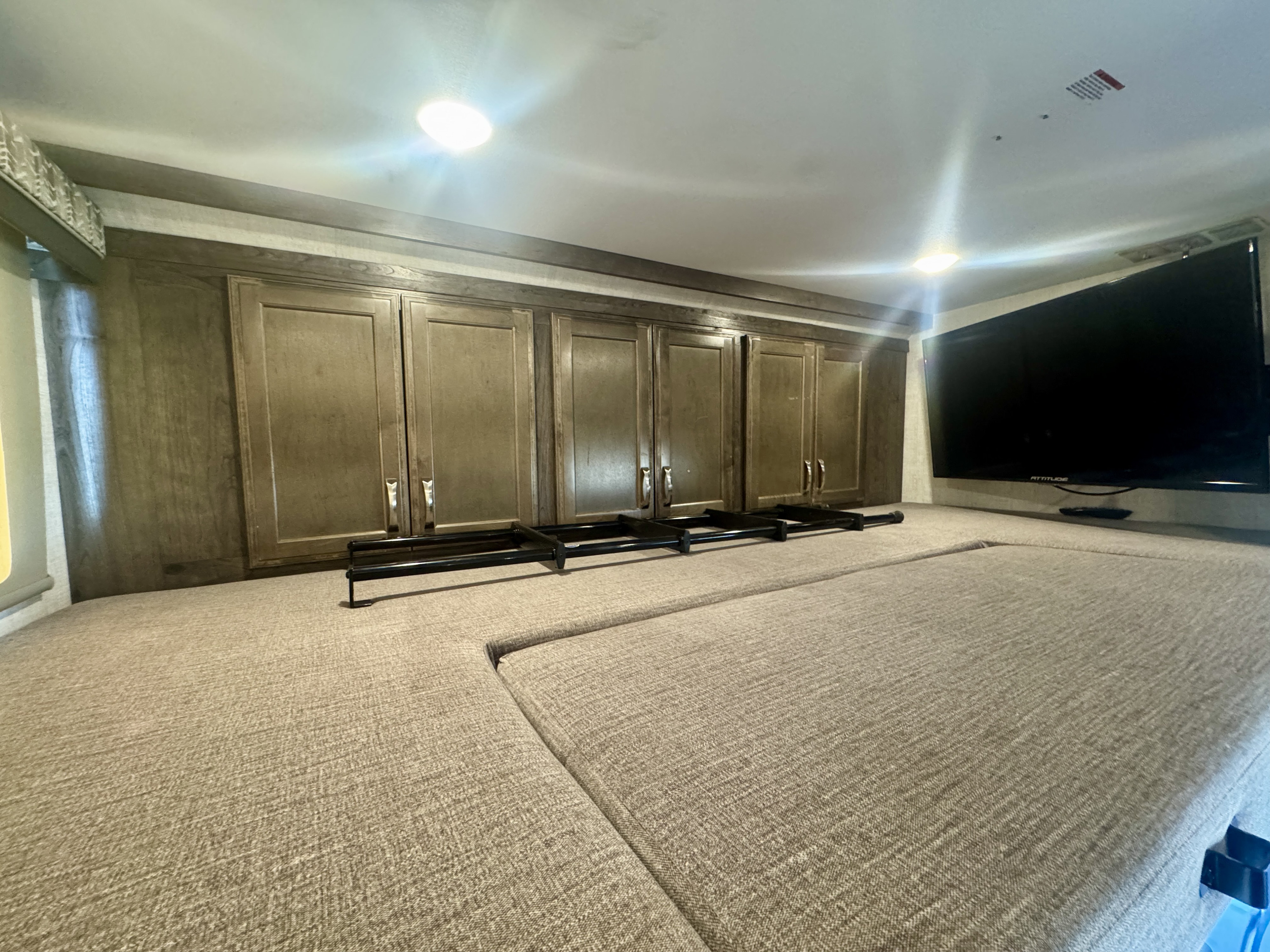 Bright RV rental bedroom: wooden overhead cabinets, wall-mounted TV, and a comfortable upholstered sleeping area with a black ladder.. Gulf Stream 6314 2023