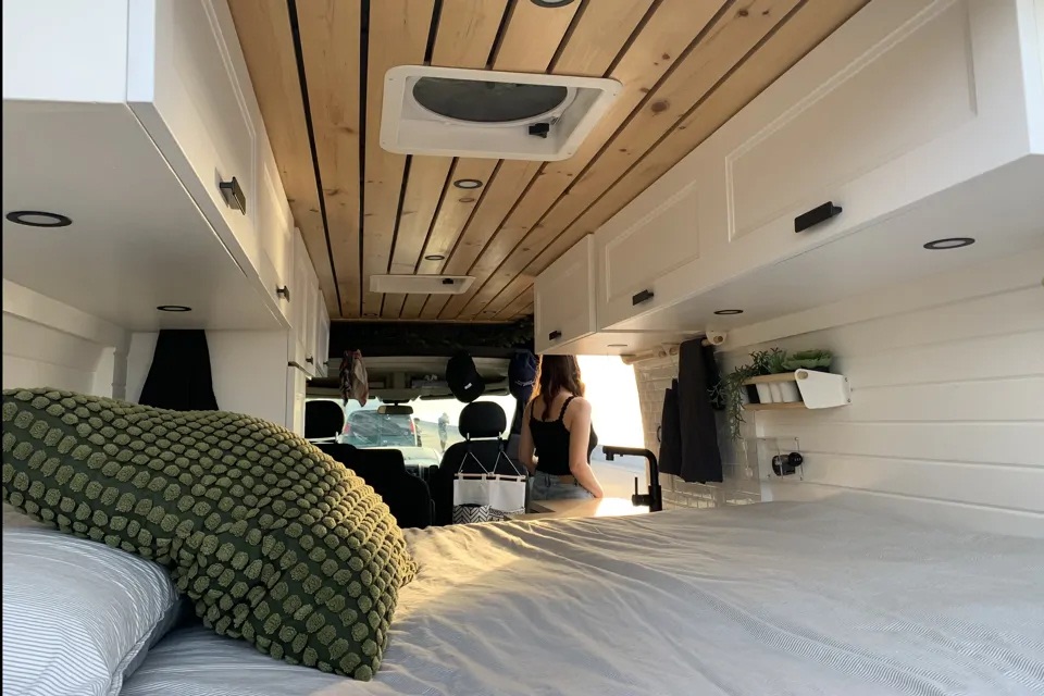 Modern RV rental interior with wooden ceiling, white cabinets, and a comfy bed. A person is seen near the kitchenette setup.. Dodge ProMaster 3500 2021