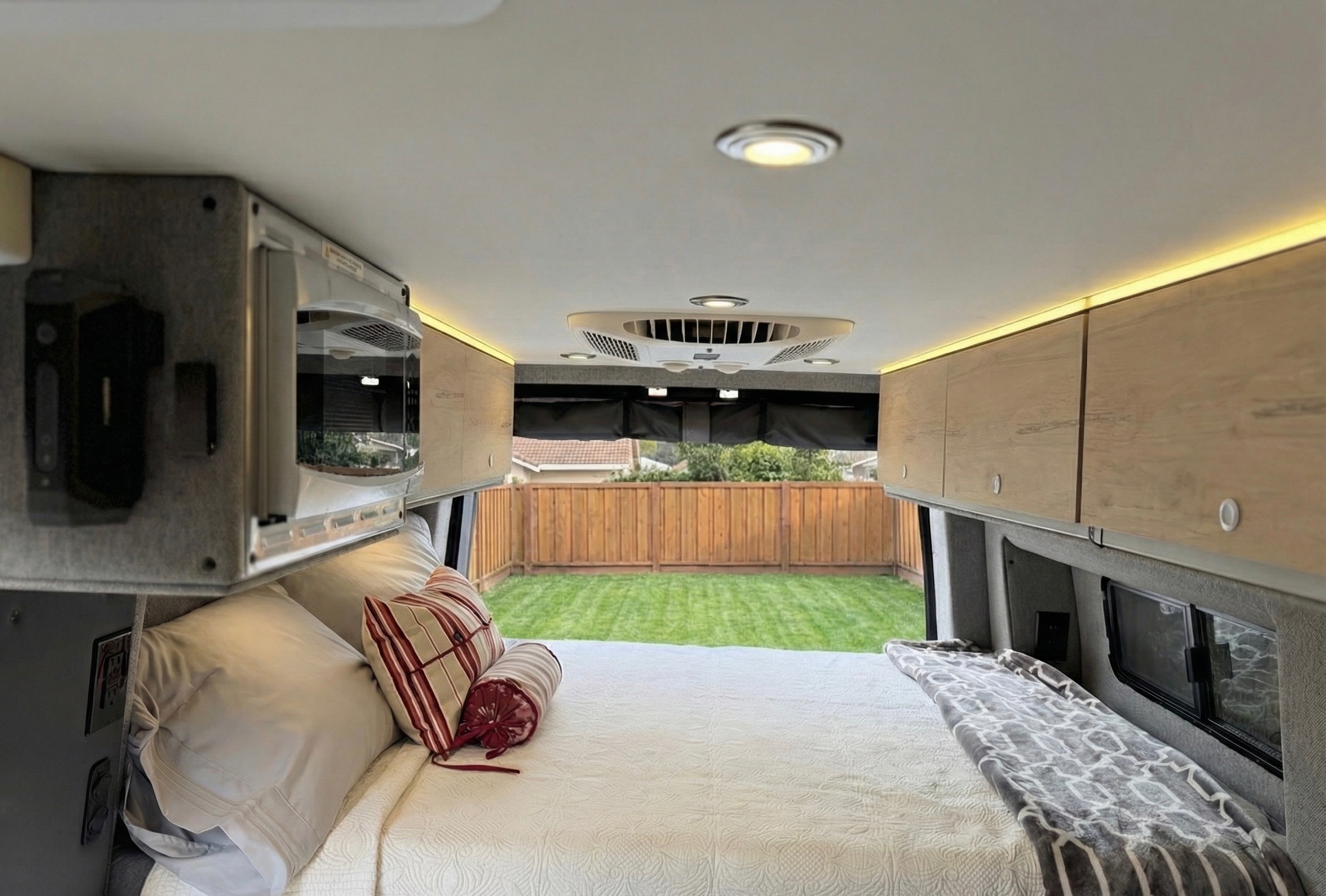 Cozy RV rental bedroom featuring a plush bed, overhead cabinets, microwave, and an open back with a green yard view for ultimate comfort.. Mercedes Sprinter Motorhome Campervan RV 2025