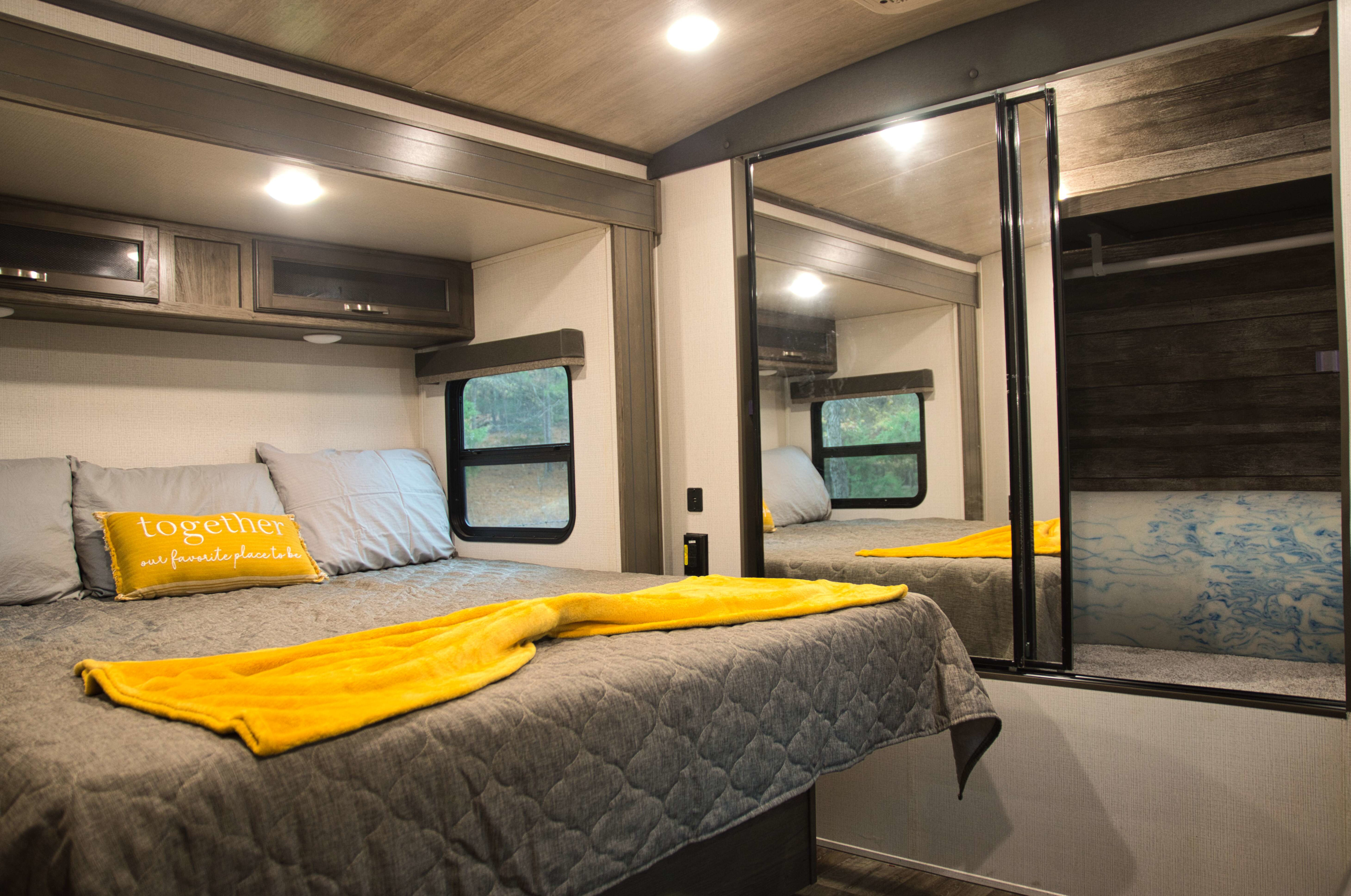 Cozy RV rental bedroom featuring a bed with grey bedding, a yellow throw, and overhead cabinets. Mirrored closet reflects the space.. Keystone RV Cougar 2022