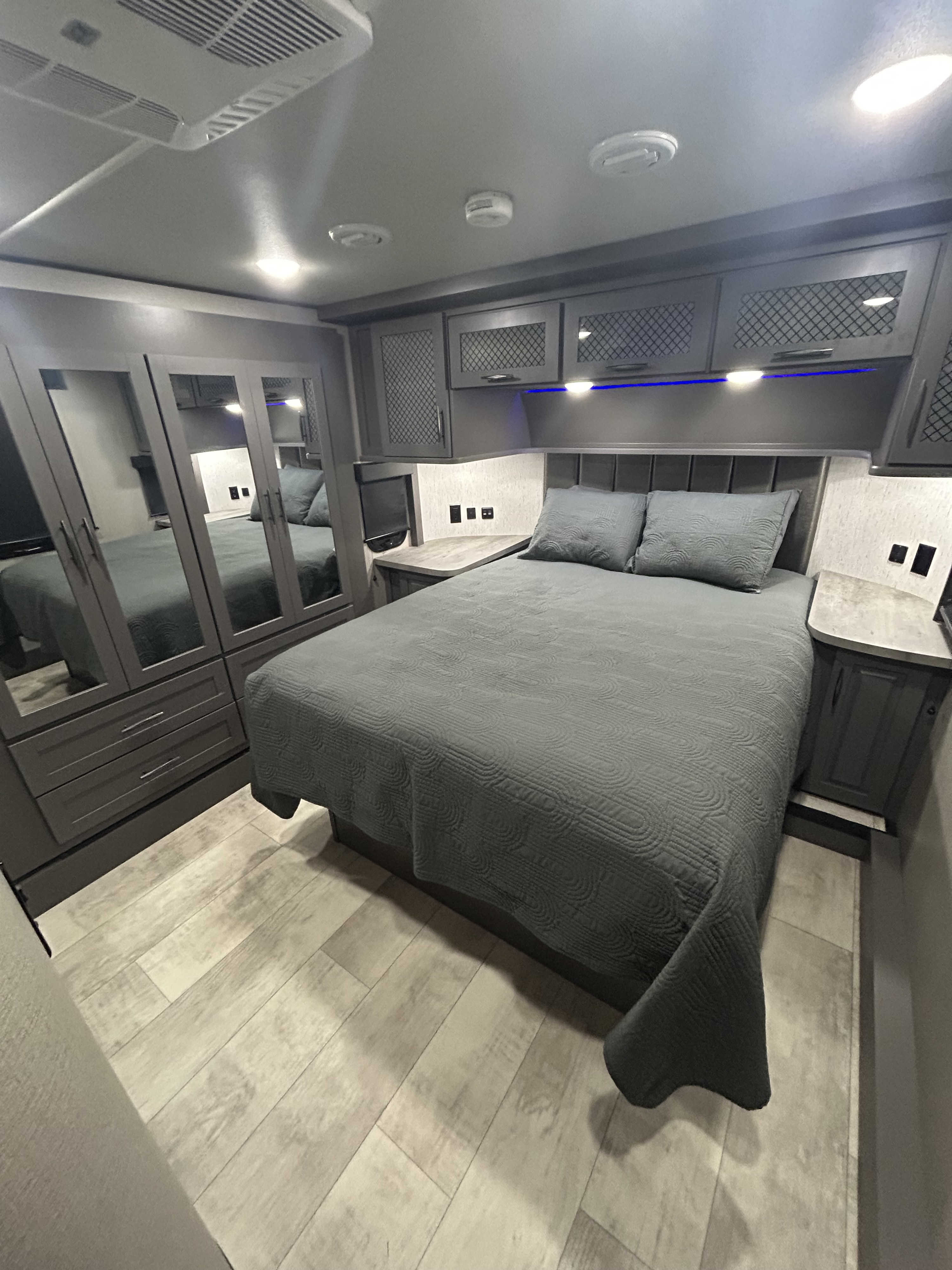Cozy RV rental bedroom with a gray quilted bed, mirrored closet, overhead cabinets, and wood-look floor. Modern design.. Grand Design Momentum 2022