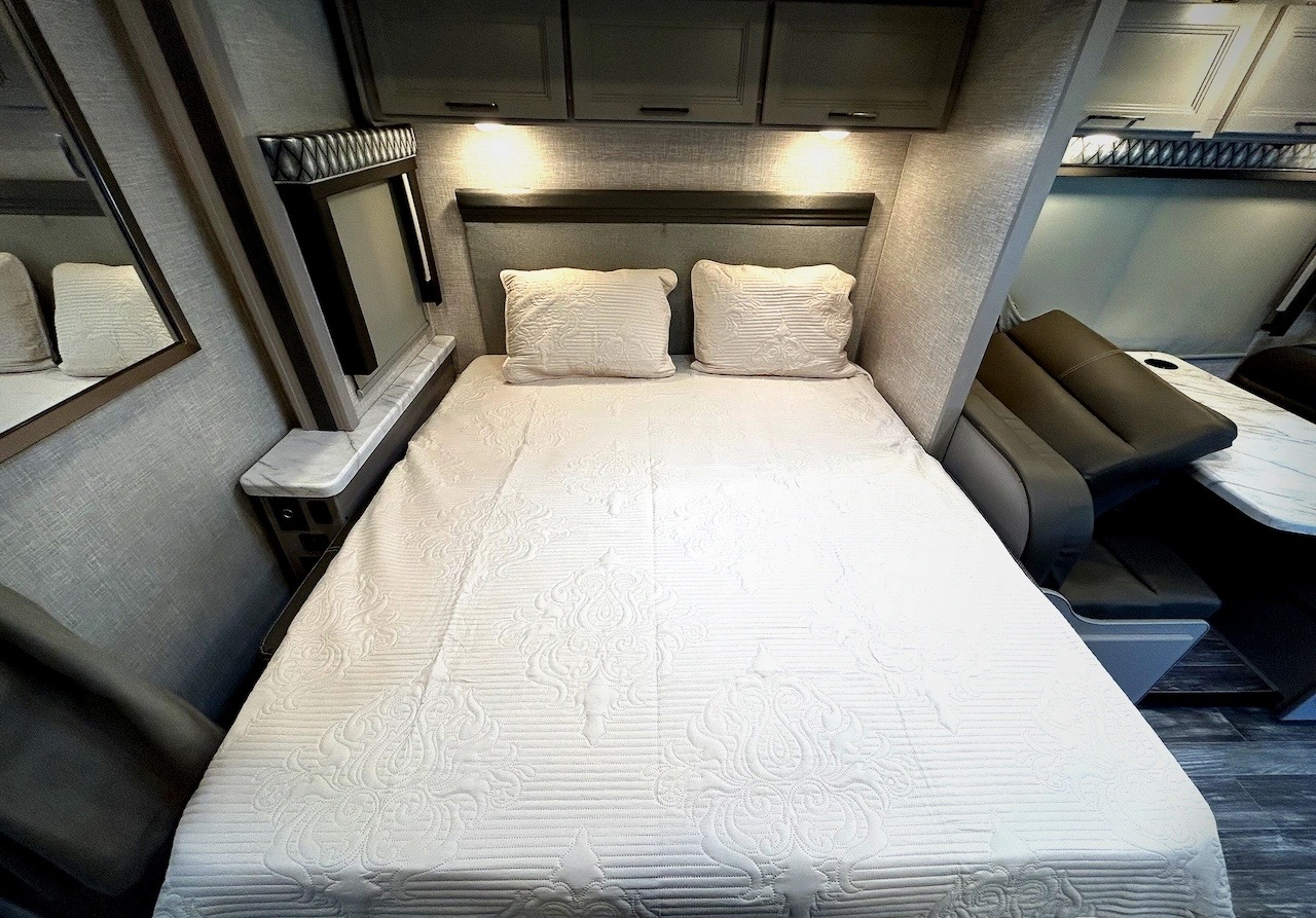 This RV rental bedroom features a comfy bed with white patterned linens, overhead cabinets, and a dinette for dining/lounge.. Thor Geneva 2023
