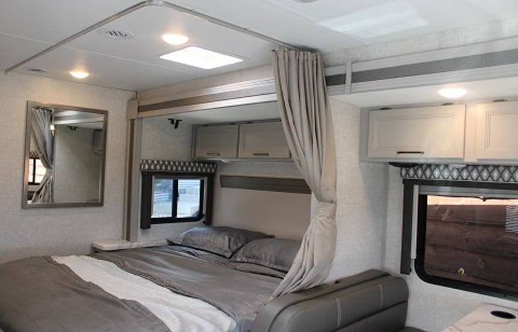 View of a modern RV rental interior, showing a comfortable bedroom with grey bedding, overhead cabinets, and natural light.. Thor Geneva 2023