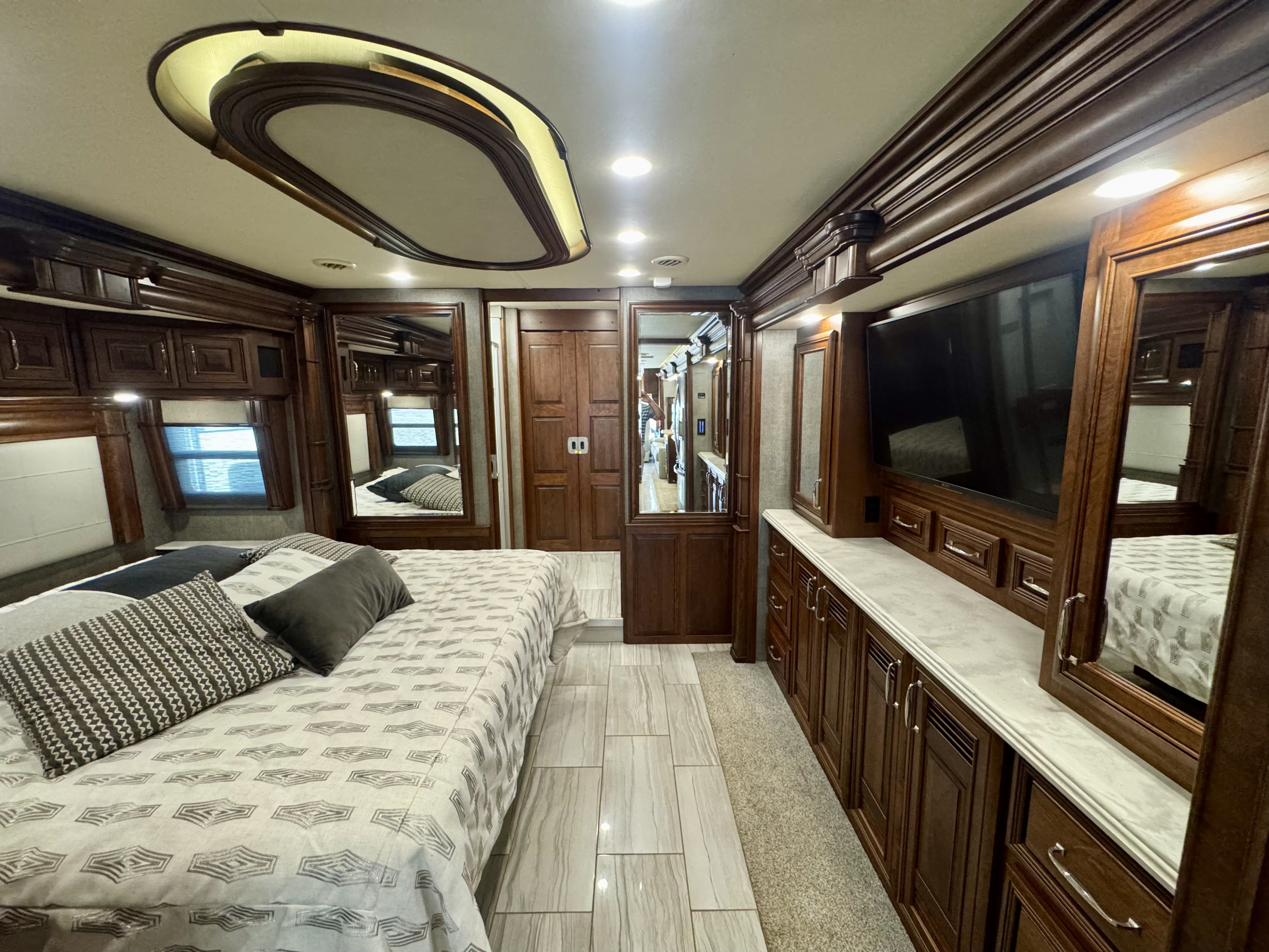 This upscale rv rental bedroom offers a comfortable bed, dark wood cabinetry, integrated TV, mirrored closets, and elegant lighting.. American Dream American Dream Motorhome 2019