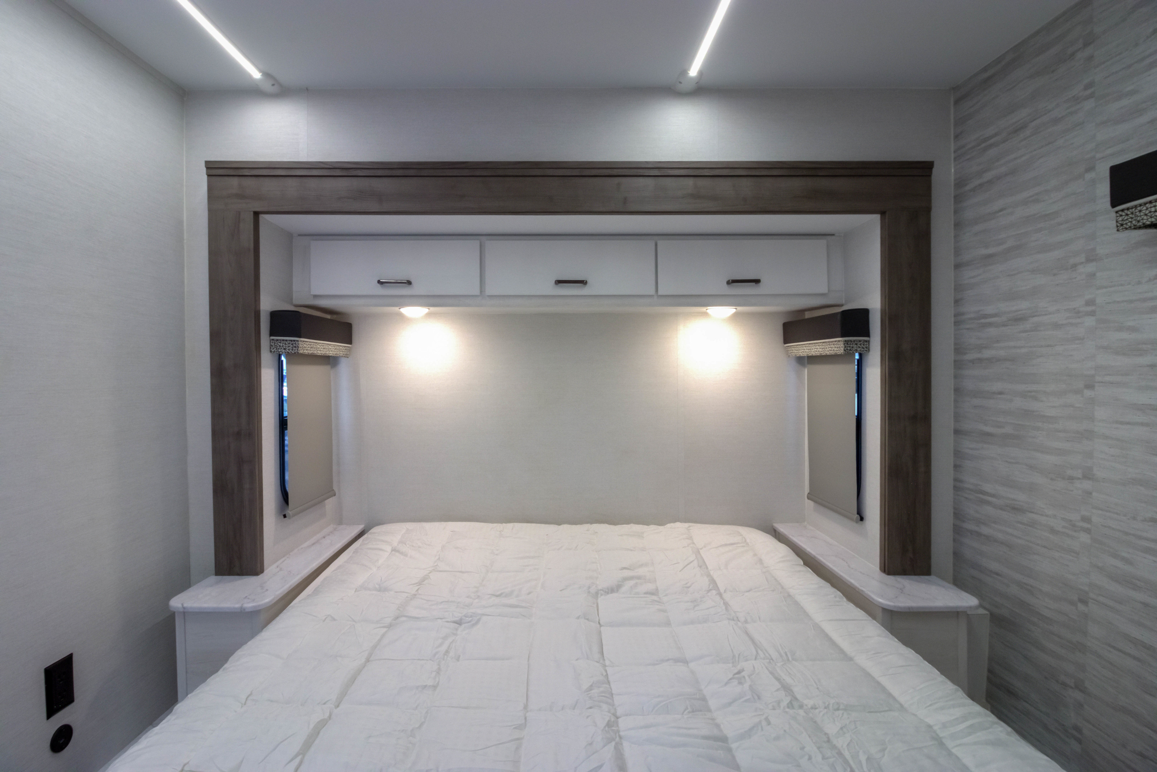 This RV rental bedroom features a white bed, overhead cabinets, side windows with blinds, and bright ceiling & bed lights.. gulf stream Conquest 2026
