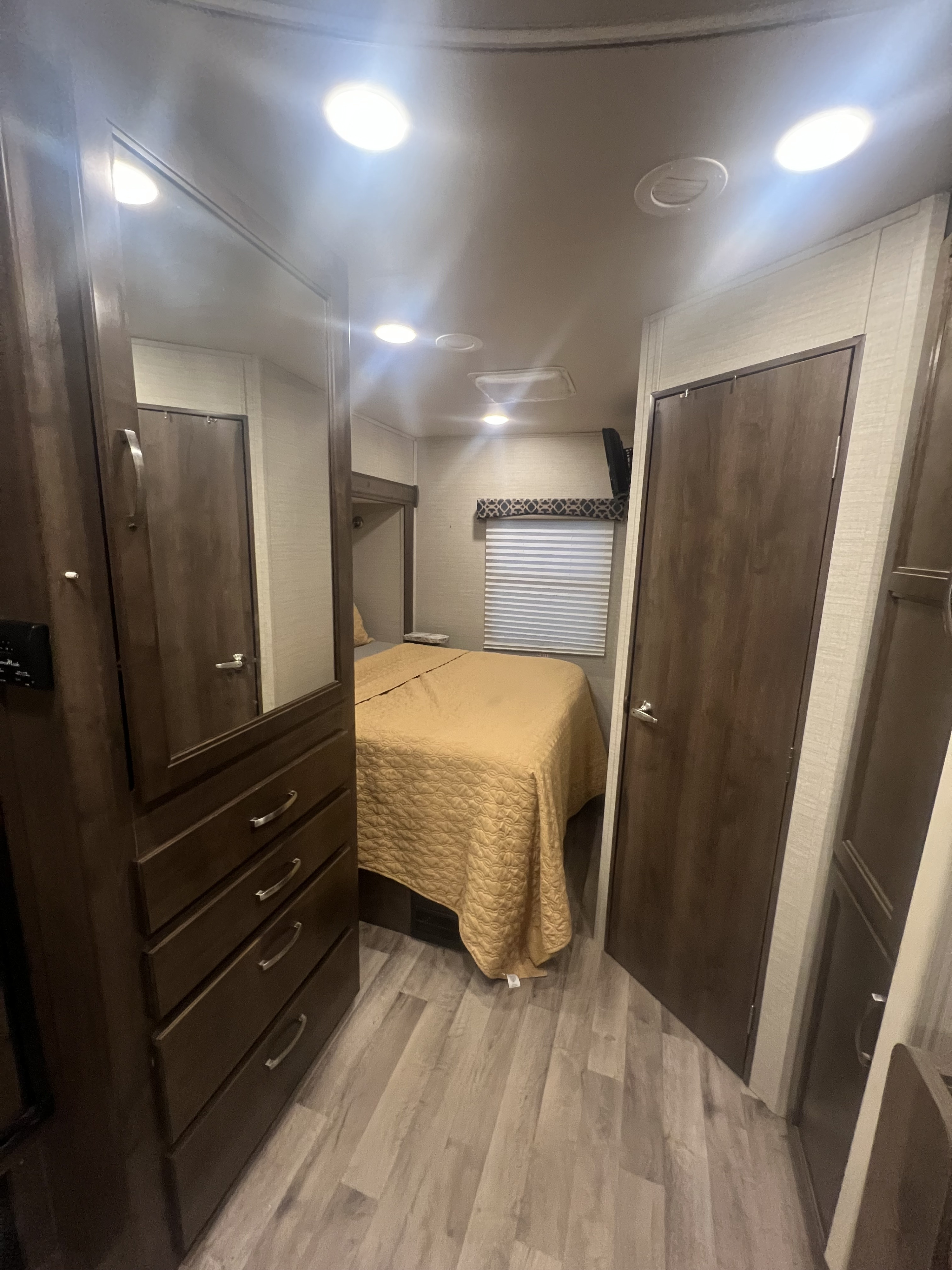 Cozy RV rental bedroom featuring a bed with a yellow quilt, wooden wardrobe with mirror, doors, window blinds, and bright lighting.. Jayco Redhawk 2019