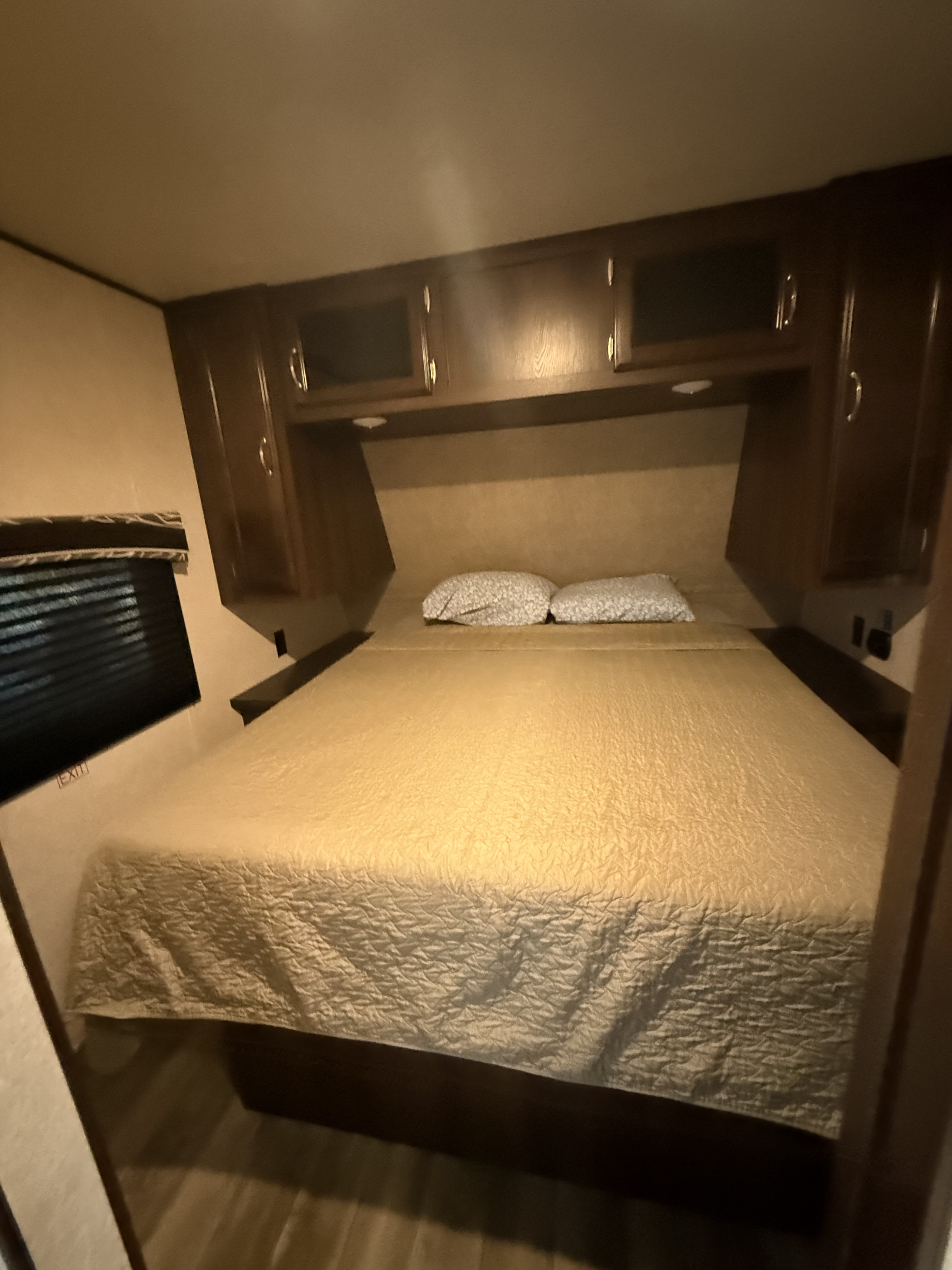 This RV rental bedroom features a comfy bed with two pillows, overhead cabinets, a window with blinds, and an "EXIT" sign.. Jayco Jay Flight 2018