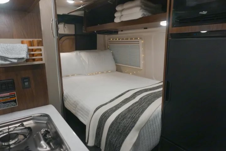This cozy rv rental features a compact bedroom with string lights, crisp white bedding, and a small kitchen area.. Winnebago Travato 2023