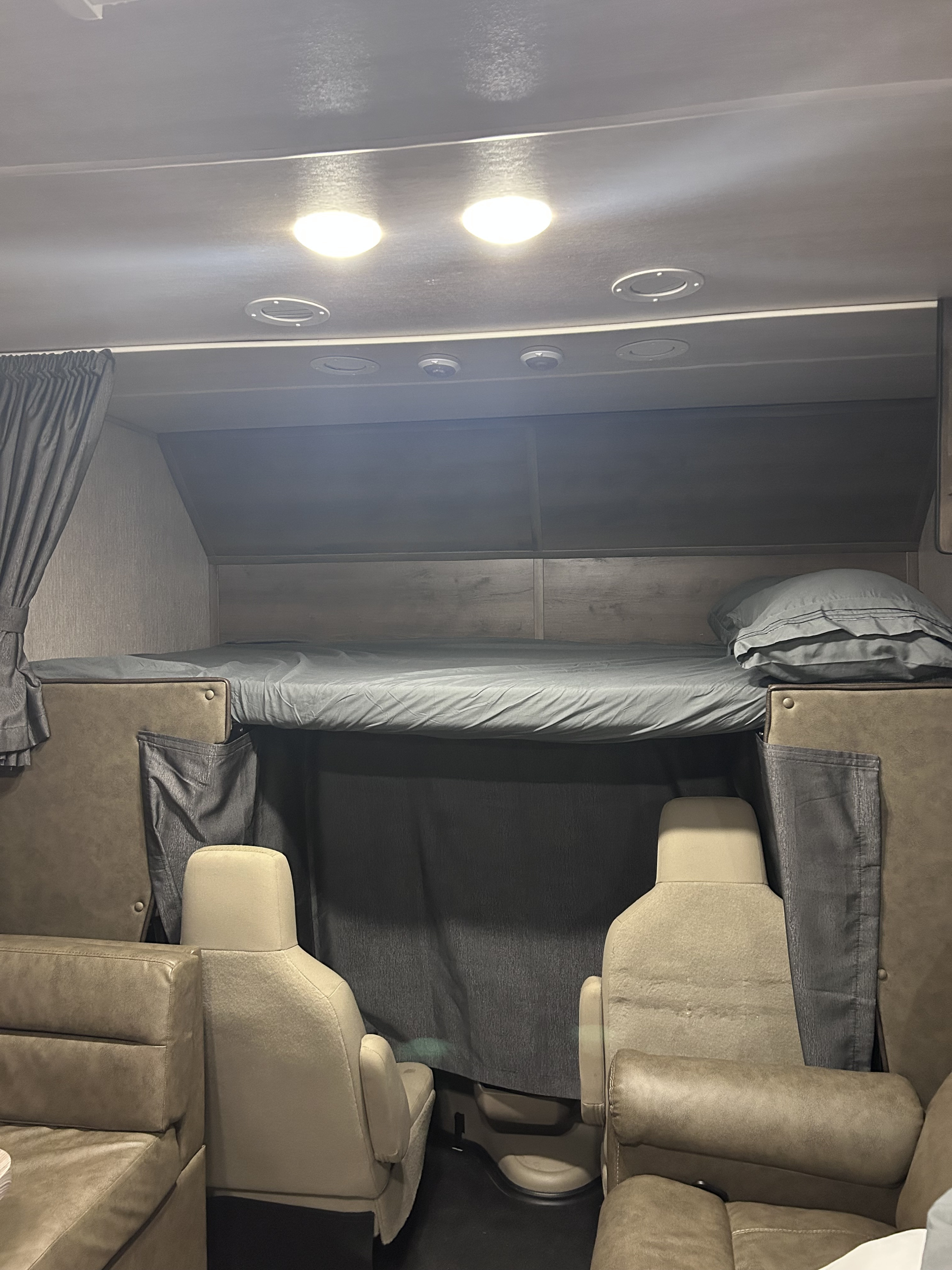 Cozy RV rental interior featuring an over-cab bunk bed with gray bedding, captain's chairs, and a sofa seating area.. Nexus RV Triumph 2022