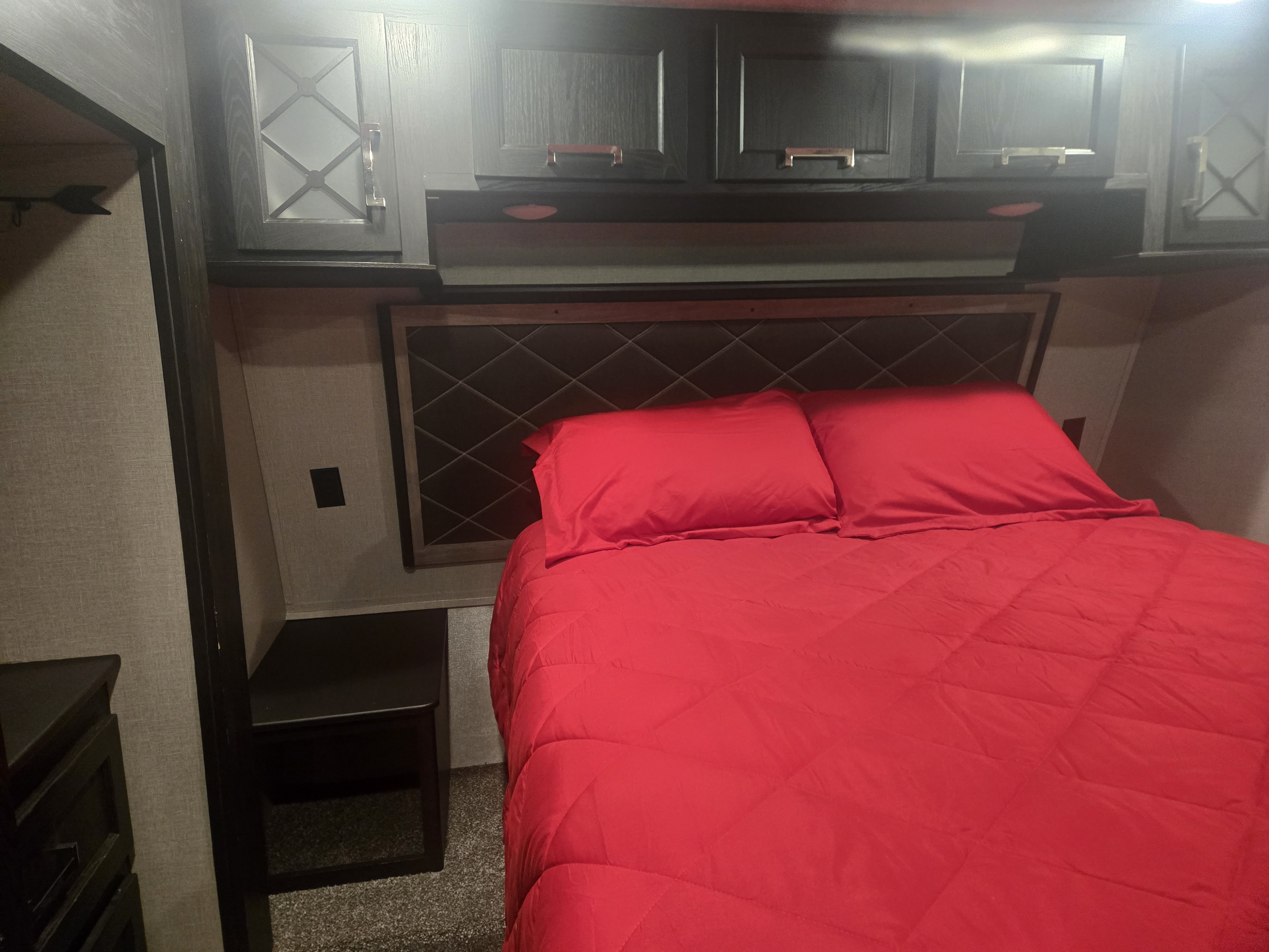 Cozy RV rental bedroom with a bed featuring bright red bedding, a diamond-patterned headboard, and dark overhead storage cabinets.. Keystone Fuzion 2018