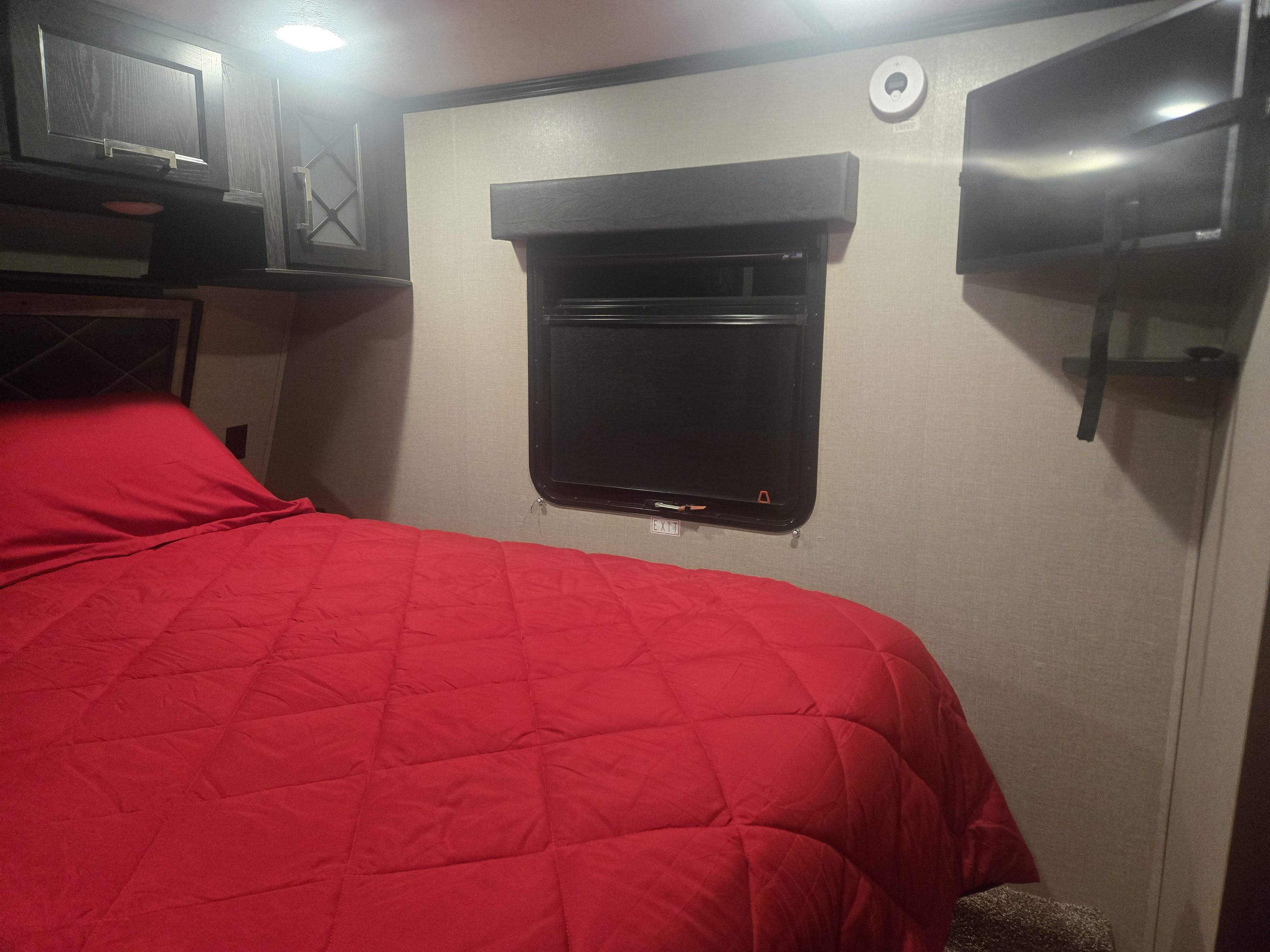 Cozy RV rental bedroom with a bed featuring bright red bedding, overhead cabinets, a mounted TV, and an emergency exit window.. Keystone Fuzion 2018