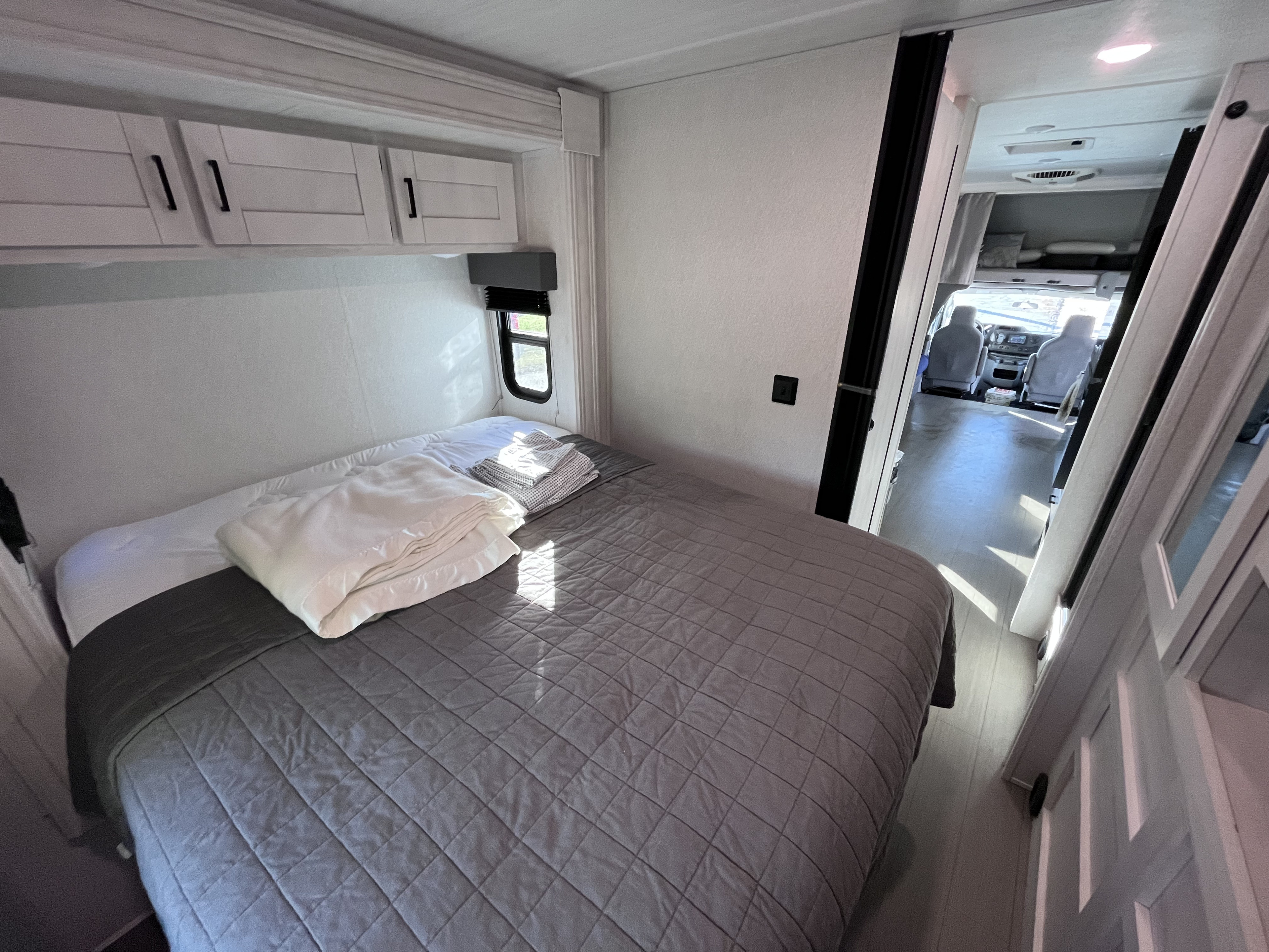 Cozy RV rental bedroom featuring a comfortable bed with a gray quilt, overhead cabinets, and a window, leading to the front cabin.. East to West Entrada 2024