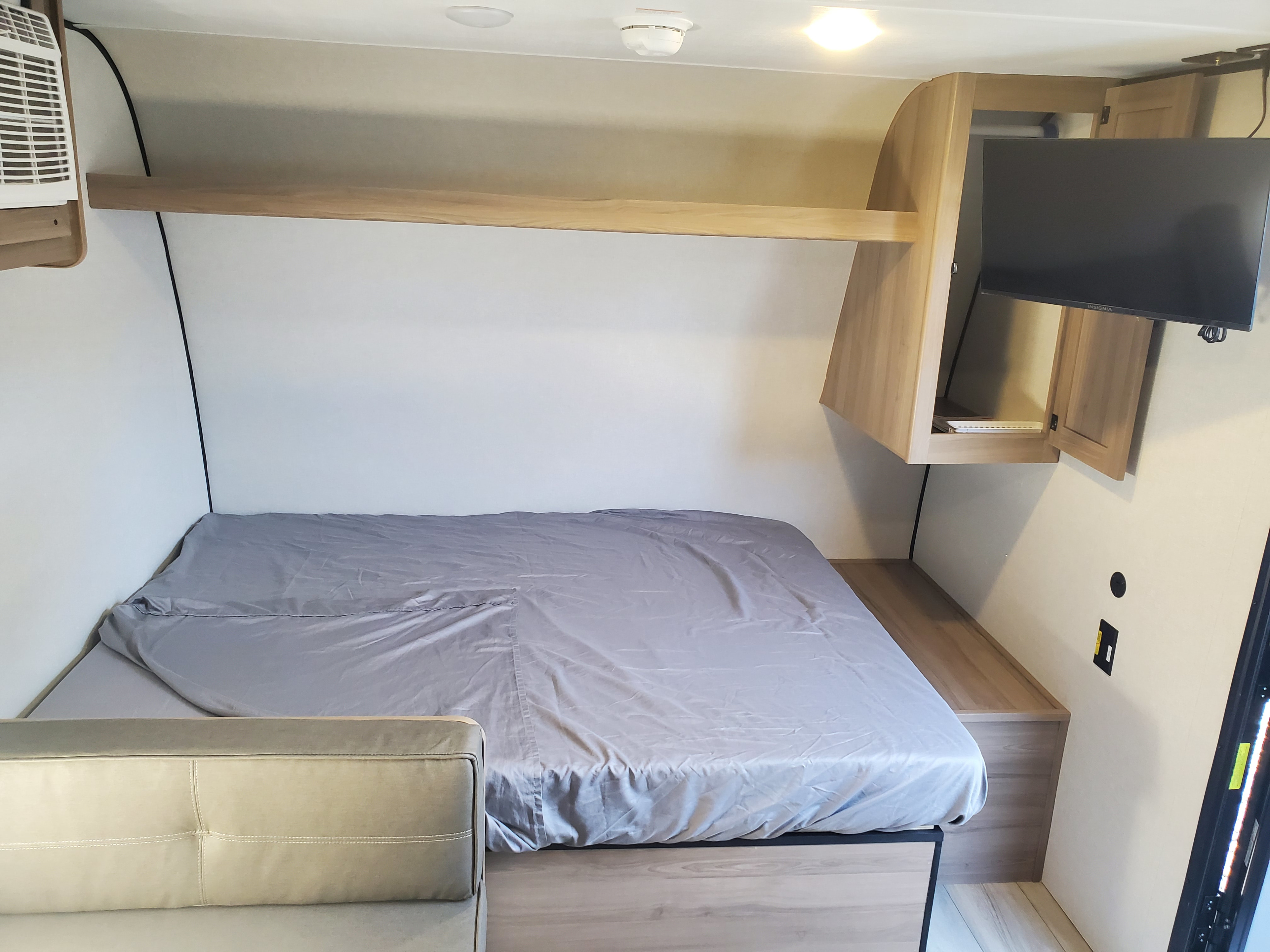 RV rental bedroom showing a bed, small sofa, wall shelf, AC unit, and TV with storage cabinets. Compact living space.. Keystone Coleman 2026