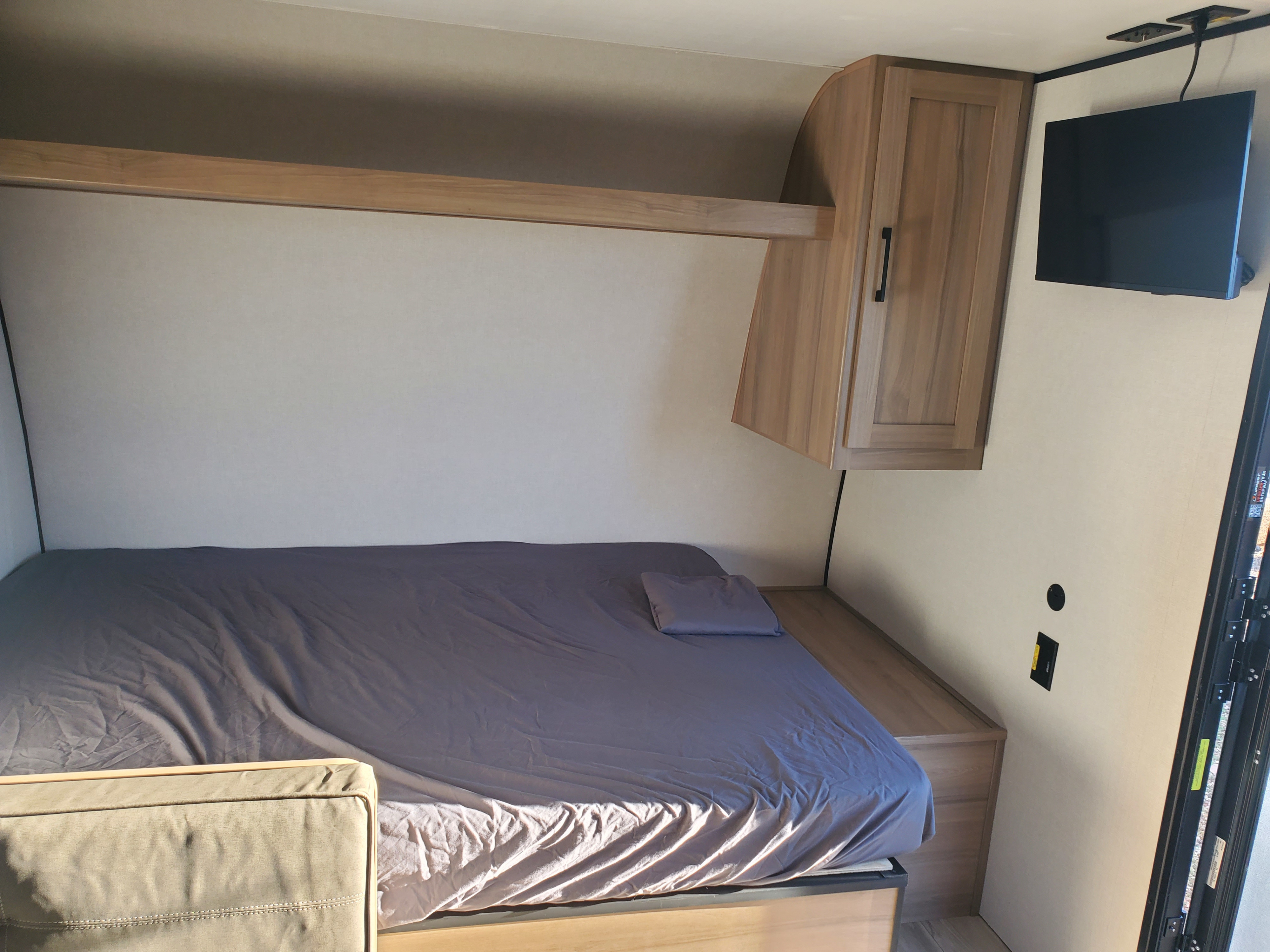 Cozy RV rental sleeping area featuring a gray-sheeted bed, overhead cabinet, shelf, and wall-mounted TV.. Keystone Coleman 2026