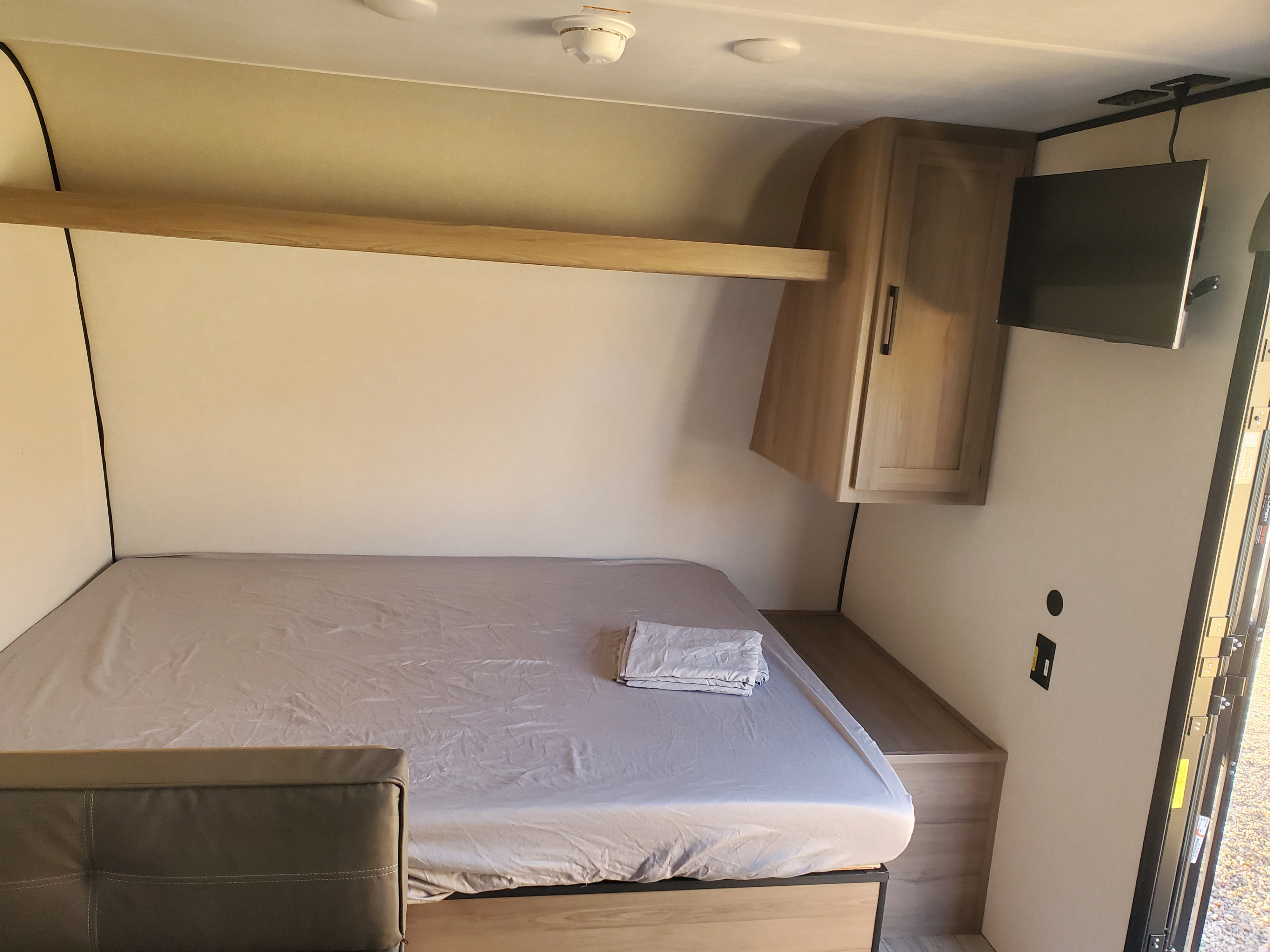 Compact RV rental bedroom featuring a fitted bed, overhead shelf, wall-mounted TV, and a storage cabinet.. Keystone RV Coleman 2026