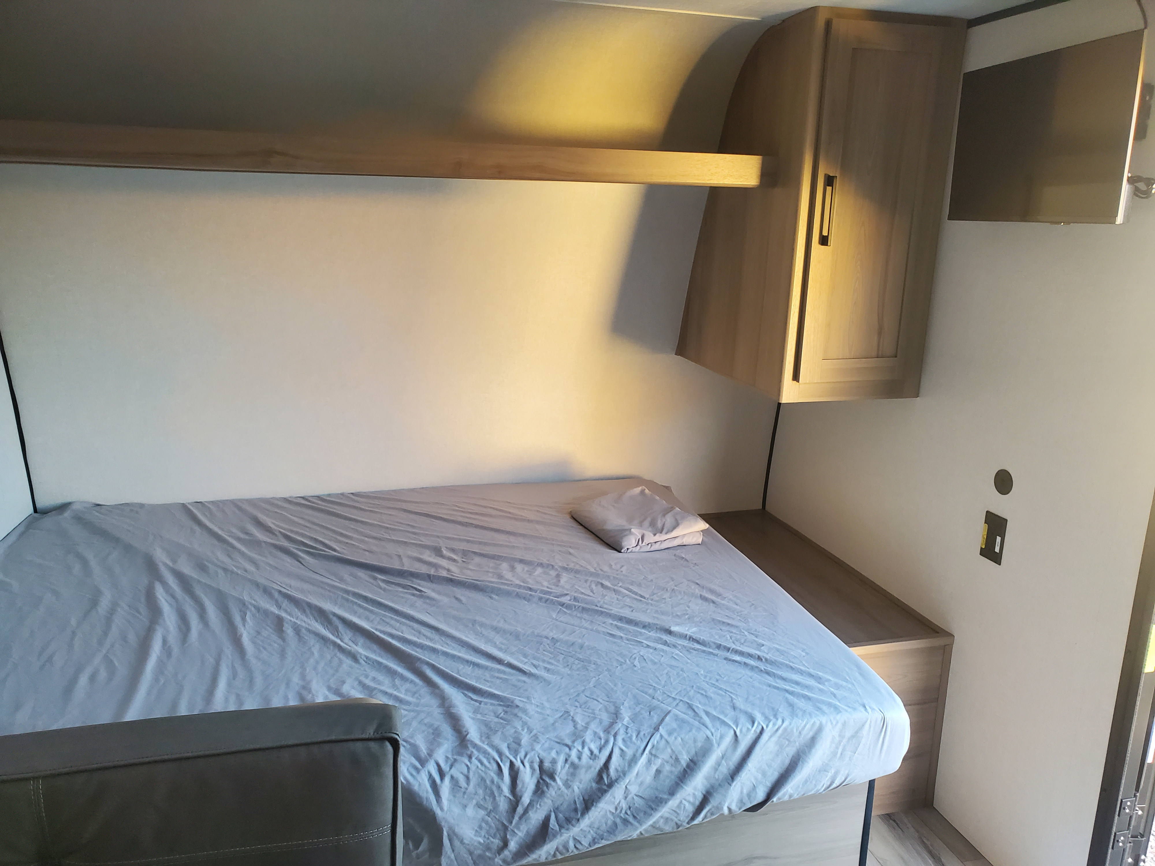 RV rental bedroom: A cozy sleeping area with a gray-sheeted bed, overhead shelf, wood cabinet, and wall-mounted TV.. Keystone Coleman 2026