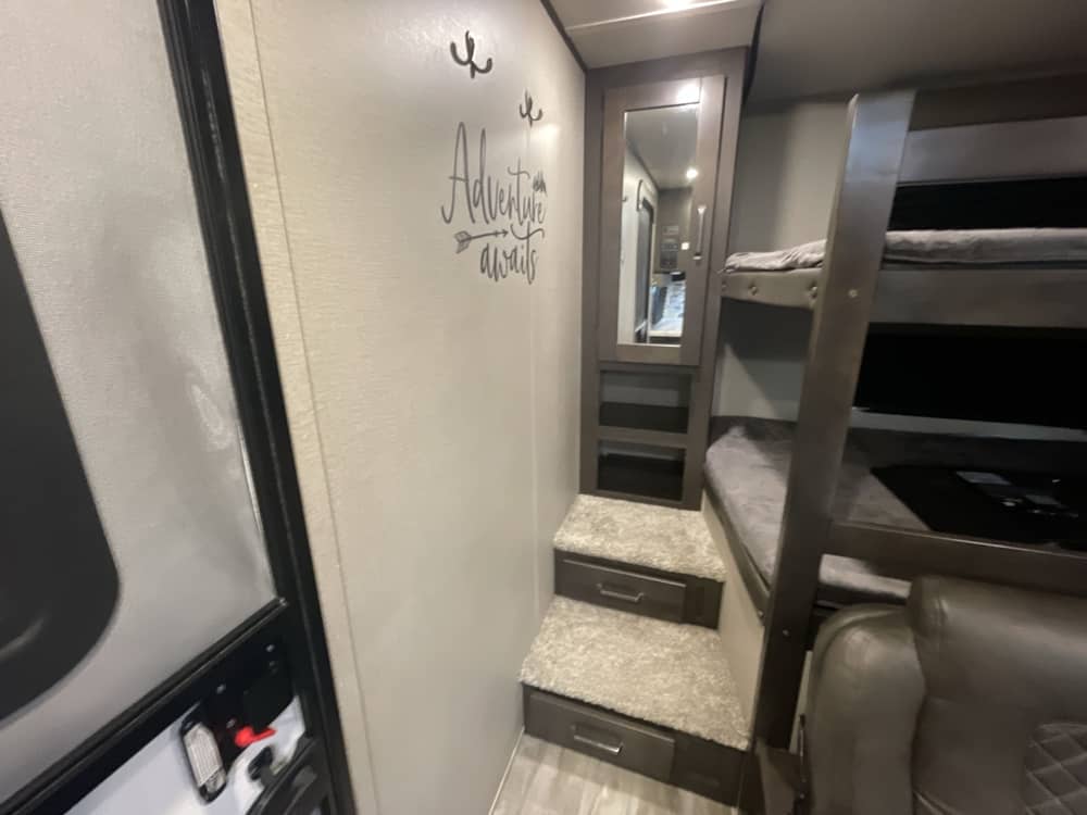 RV rental bunk room: stacked beds, stairs with storage drawers, mirrored cabinet, and 'Adventure awaits' wall art.. Grand Design Transcend Xplor 2022