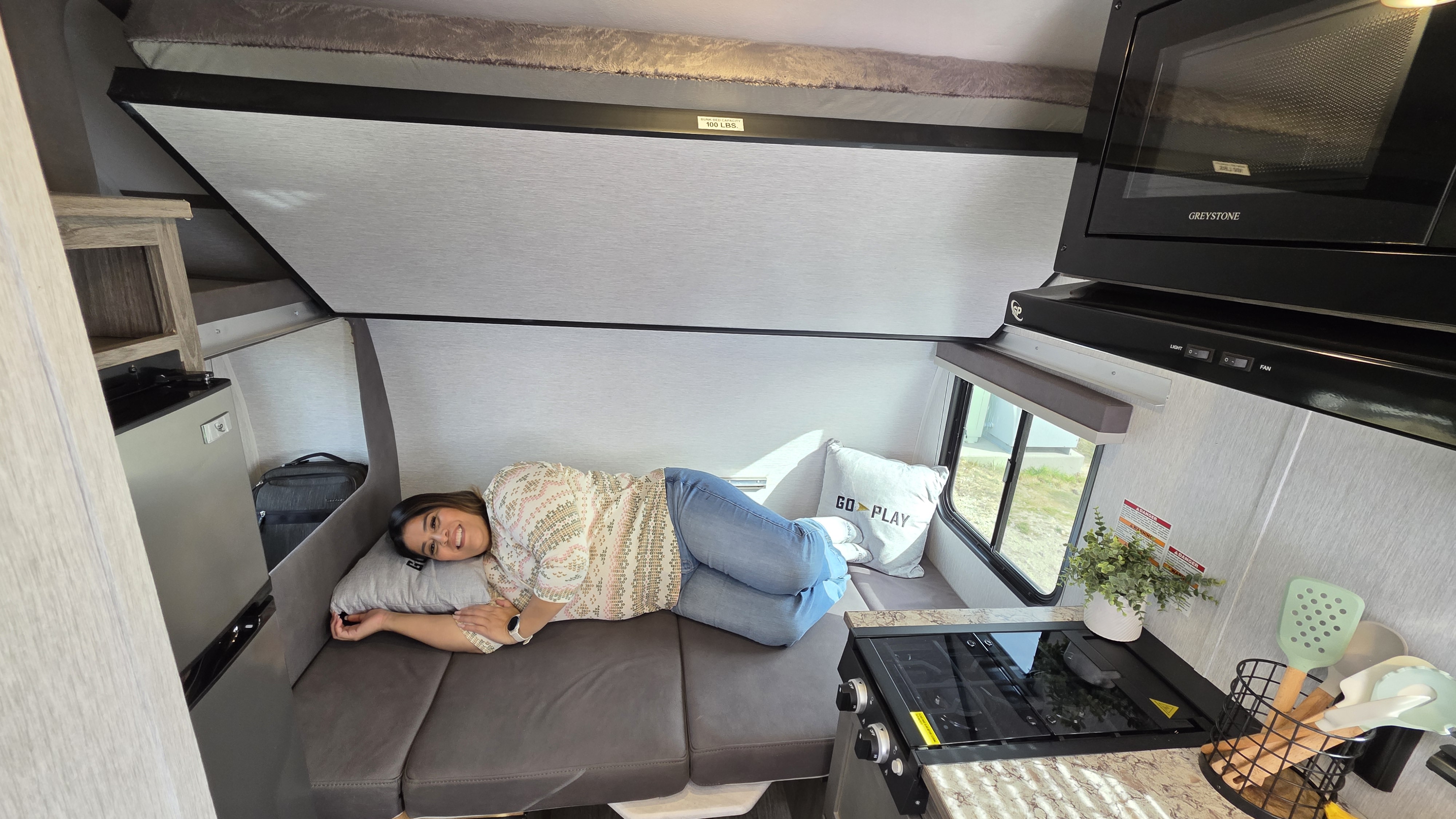 Woman relaxes inside a cozy RV rental, featuring a compact kitchen, microwave, and upper bunk bed.. Wayfinder RV Go Lite 2025