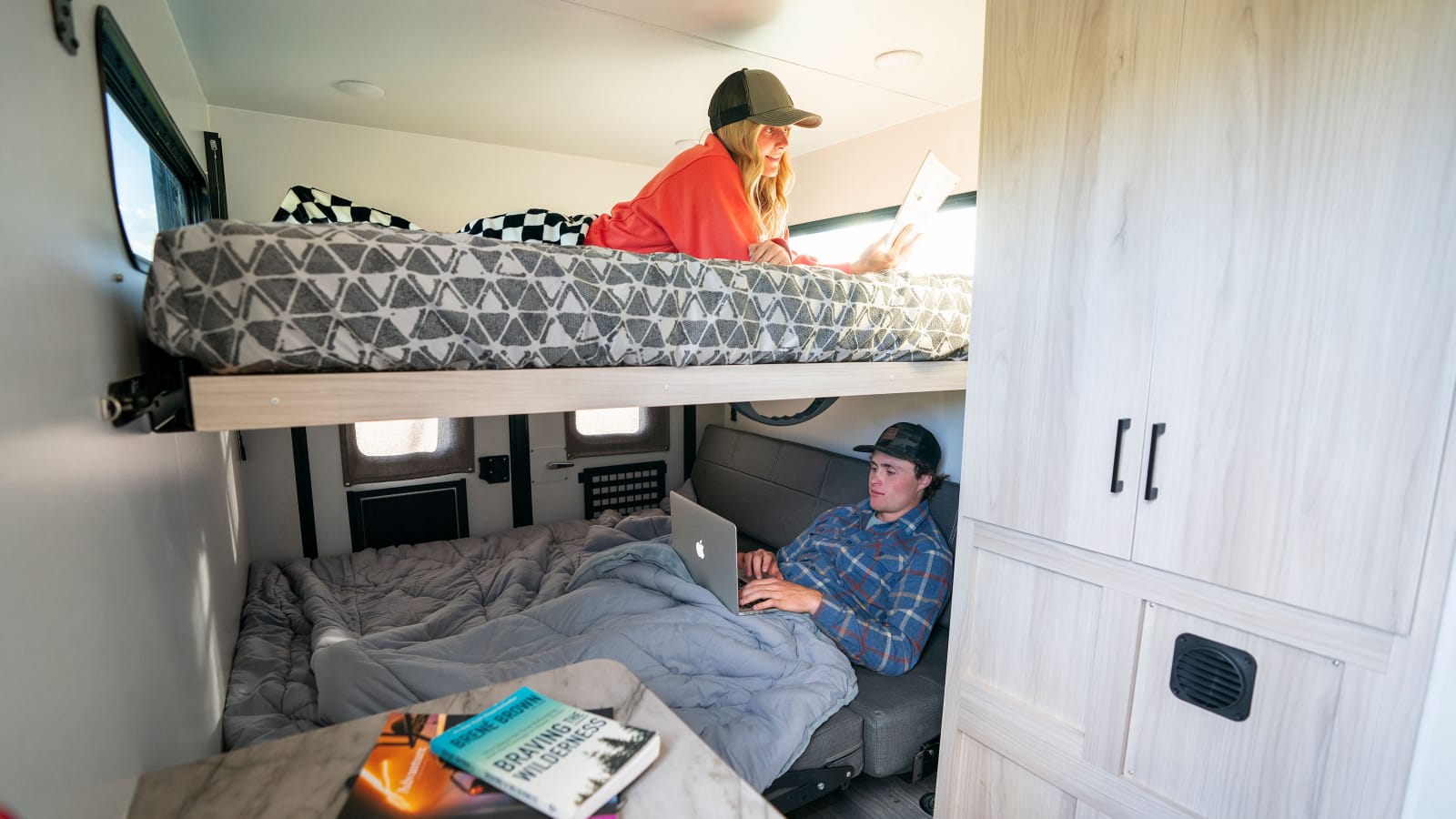 Inside an RV rental, two people relax in comfortable bunk beds. A woman reads on the top bunk, while a man uses a laptop below.. Winnebago Hike 100 2023