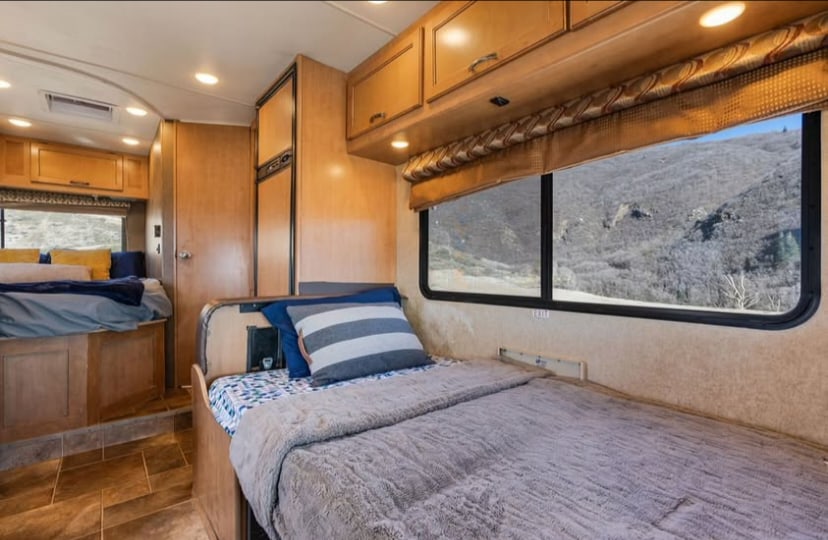 This cozy RV rental offers a comfy bedroom with two beds, warm wood tones, and a large window framing a serene mountain view.. Thor Four Winds 2017