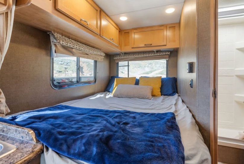 RV rental bedroom features a cozy bed with a blue blanket, overhead storage, window views, and an adjacent shower.. Thor Four Winds 2017